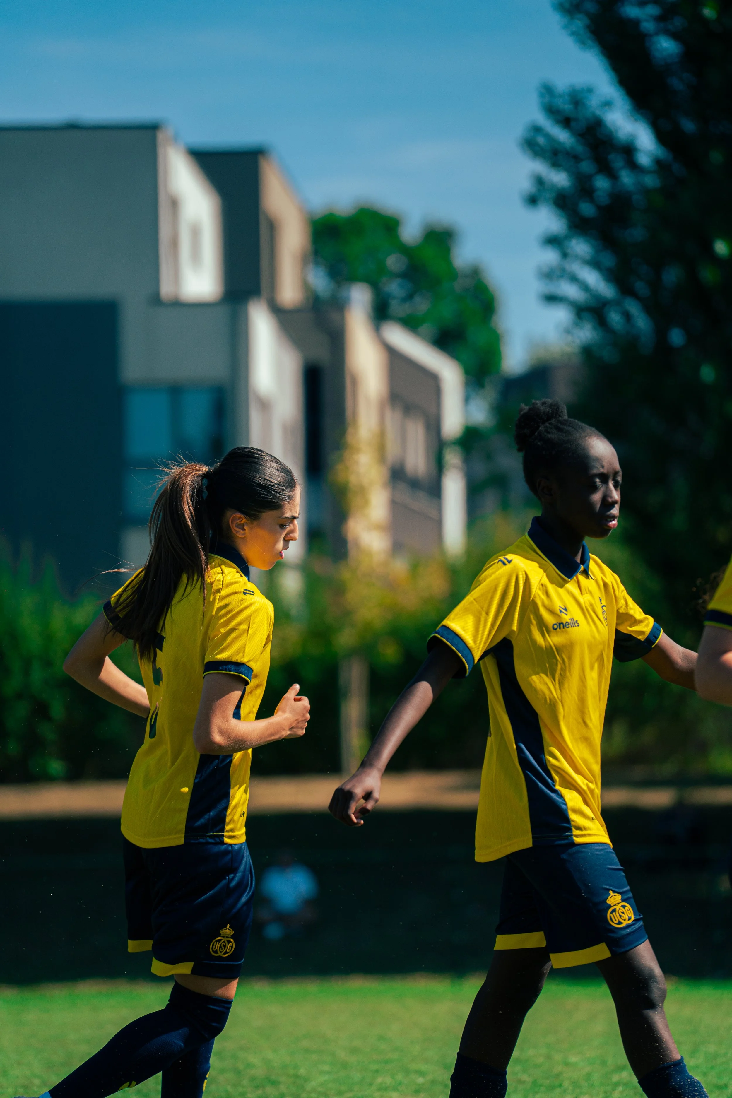 Two female soccer players wearing yellow and blue uniforms jogging on a soccer field with trees and residential buildings in the background.