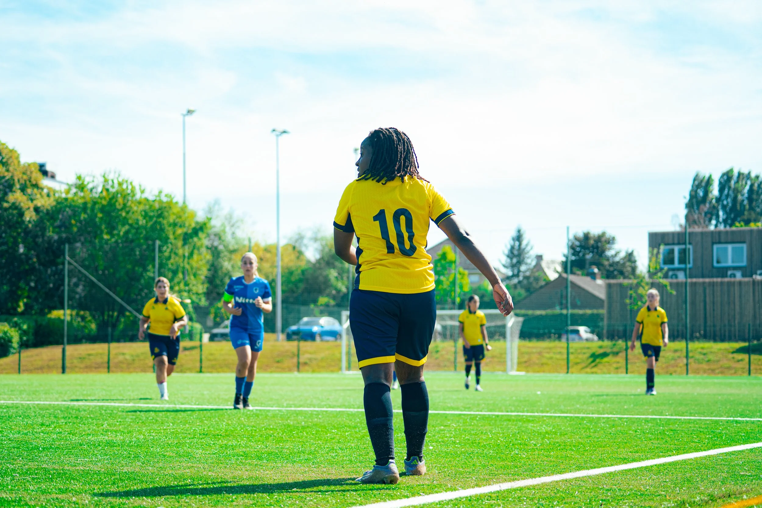 A female soccer player with dreadlocks wearing a yellow jersey with the number 10 stands on a soccer field, with other players in yellow and blue jerseys nearby, trees, houses, and a blue sky in the background.