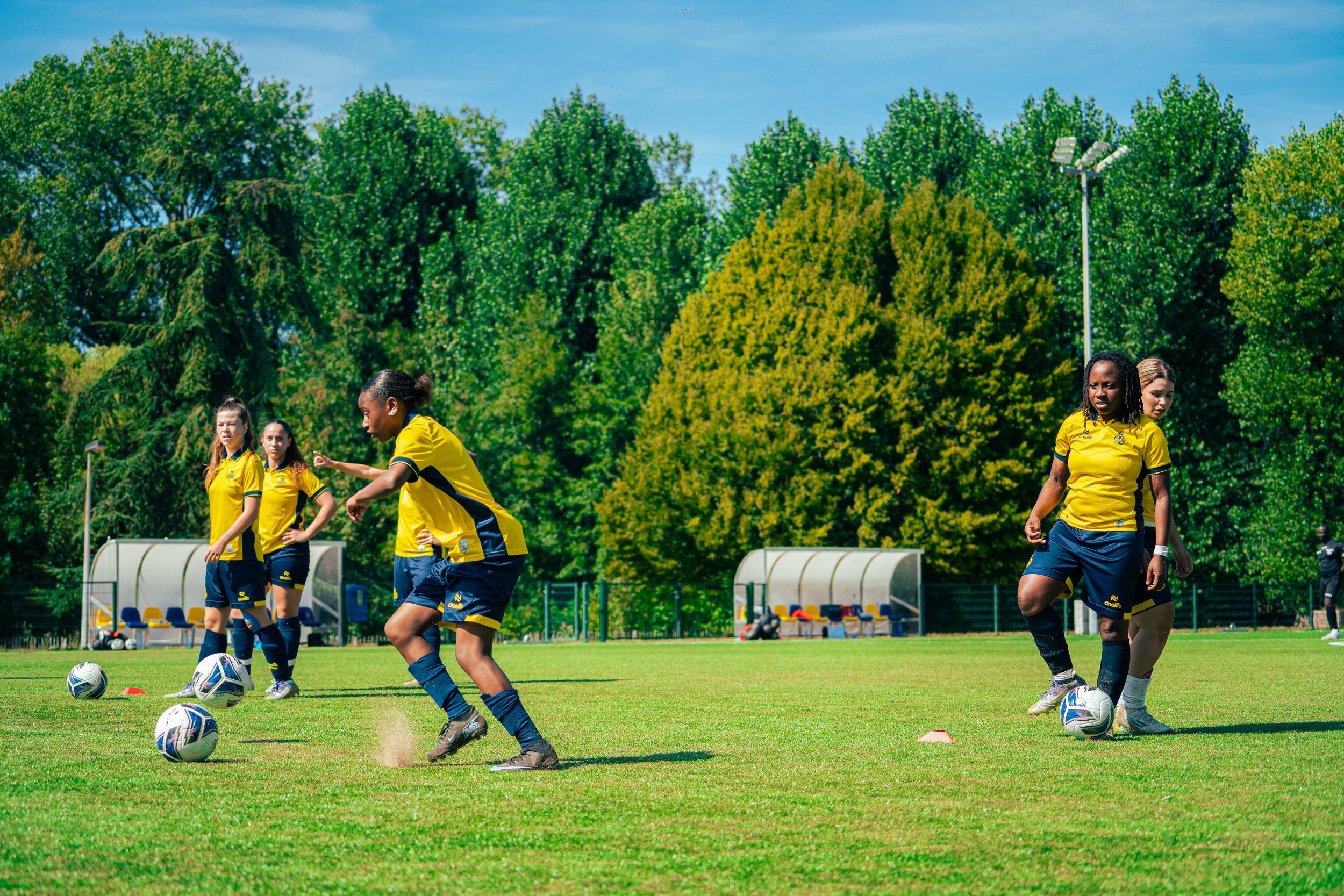 Women soccer players in yellow and navy uniforms on a field practicing with soccer balls on a bright, sunny day surrounded by green trees.