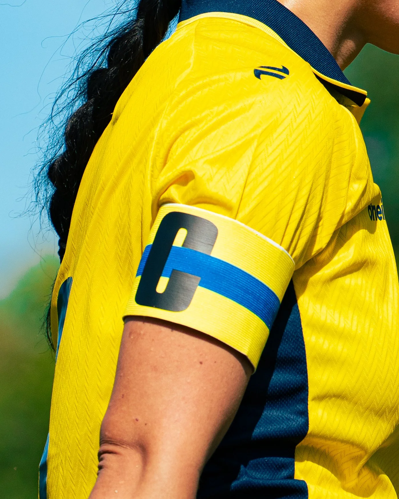 Close-up of a person wearing a yellow and blue sports jersey, with a blue and yellow captain's armband on their arm, and their dark hair in a braid.