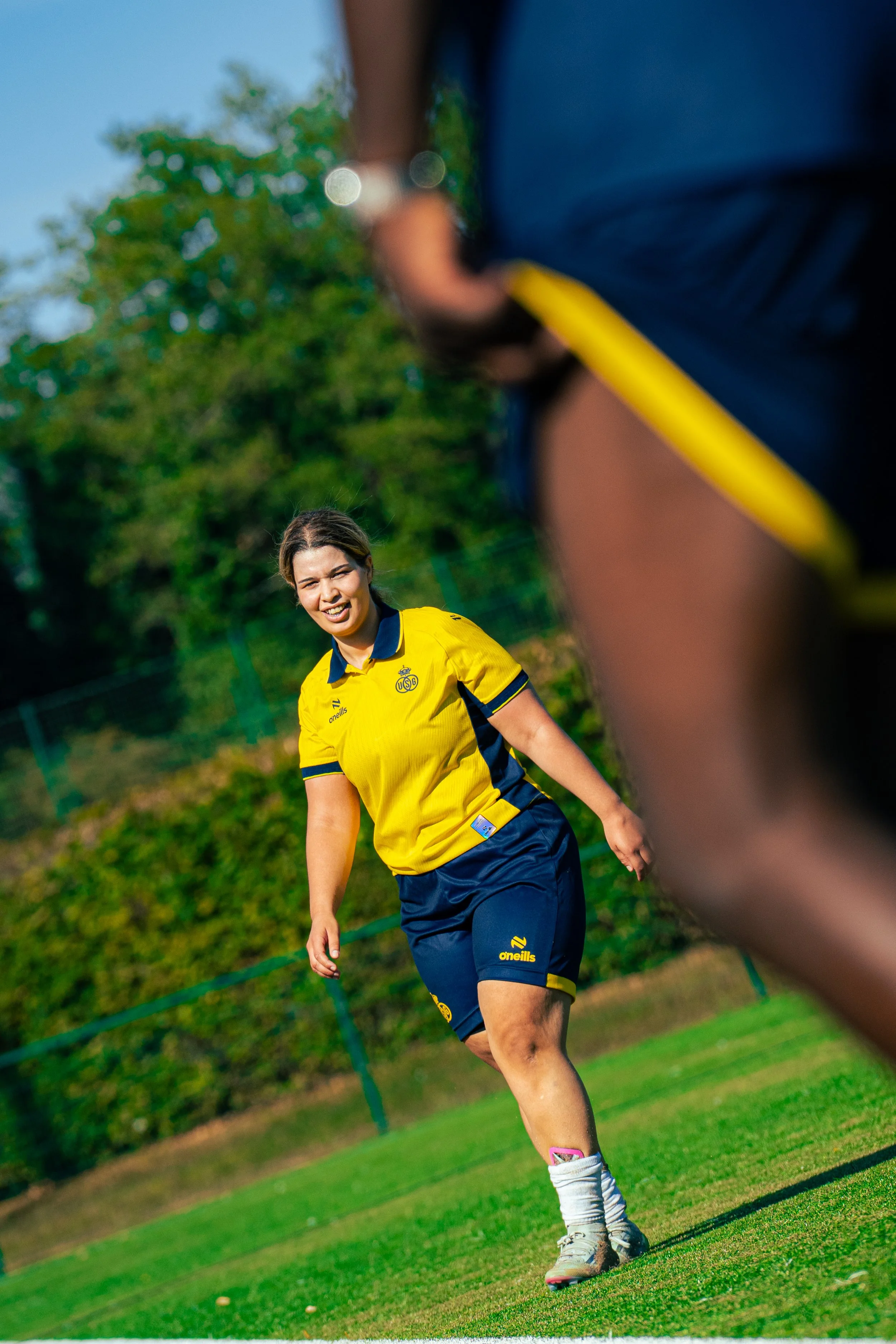 A female golfer in a yellow and navy golf uniform on a green golf course, smiling and looking at the camera, with trees in the background.