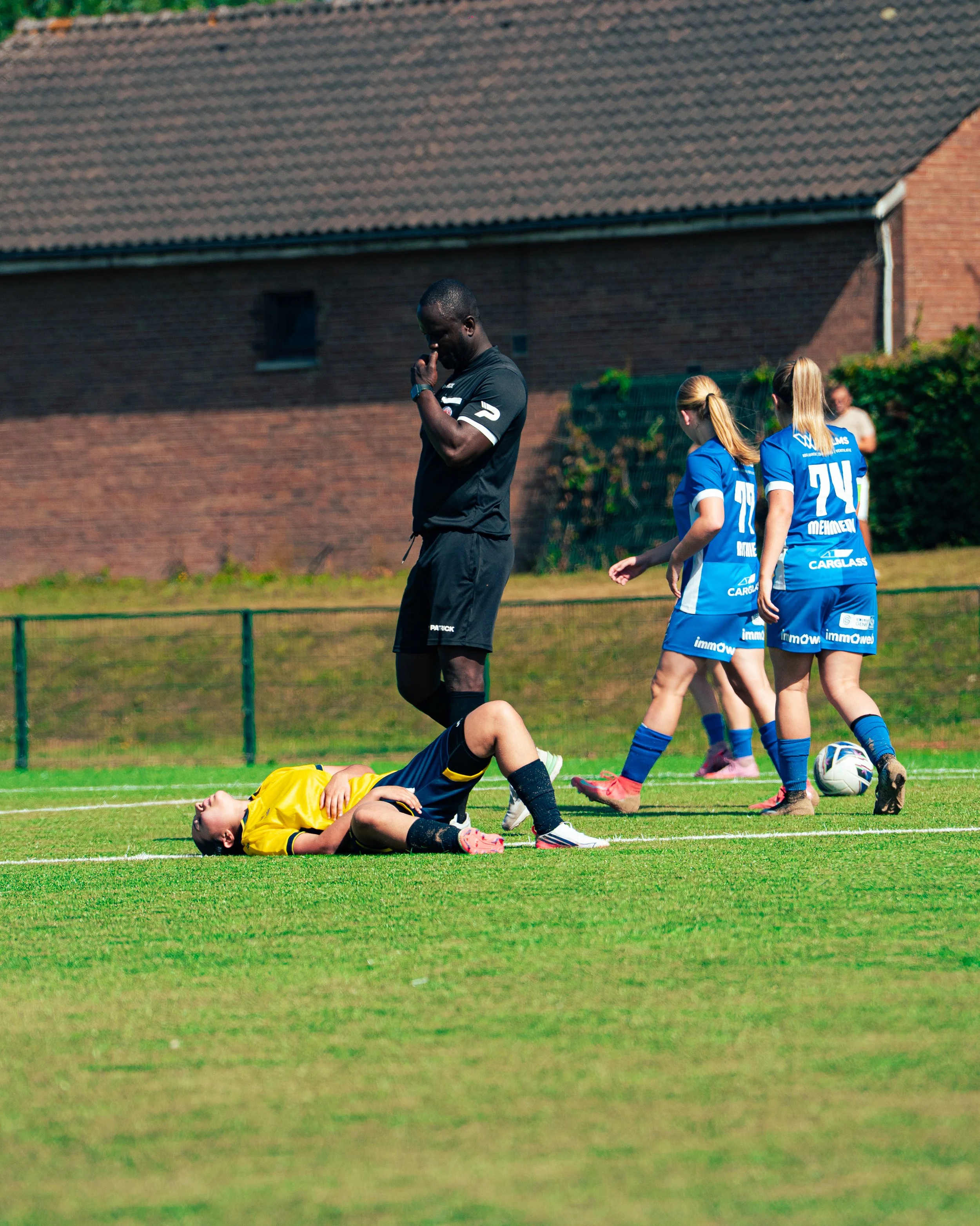 A soccer player in a yellow jersey lies on the ground holding his stomach with his hand, as two female players in blue jerseys walk nearby on a grassy field. An official stands with his head bowed and one hand touching his face, appearing concerned. 