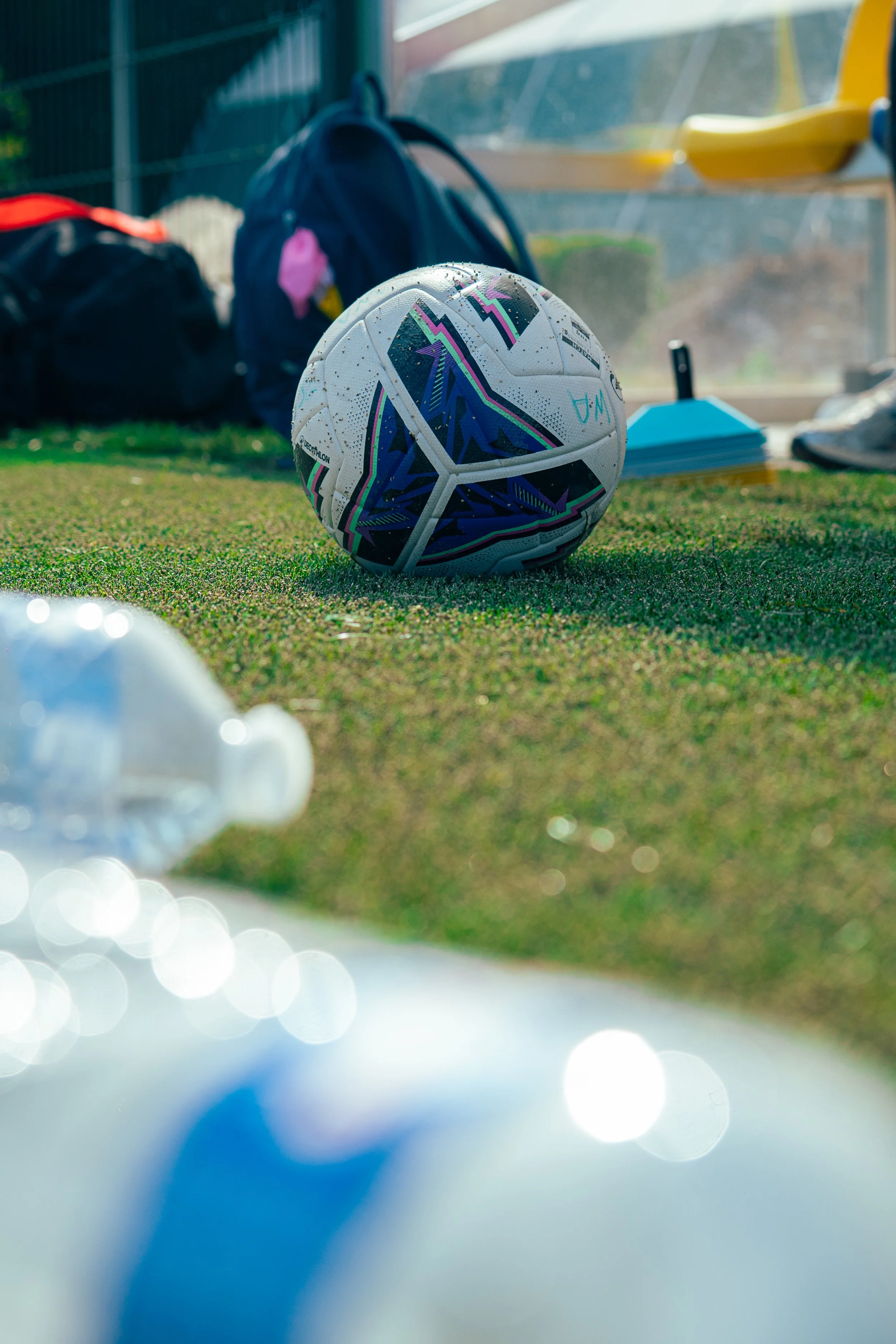 Close-up of a soccer ball on the ground with a bench, backpacks, and spray bottles in the background, suggesting a sports practice or game setting.