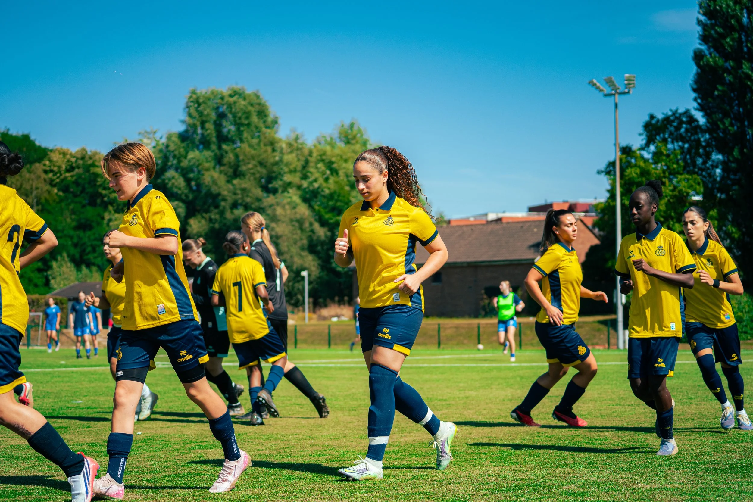 A group of women in yellow and blue soccer uniforms practicing on a green field under a clear blue sky, with trees and sports lights in the background.