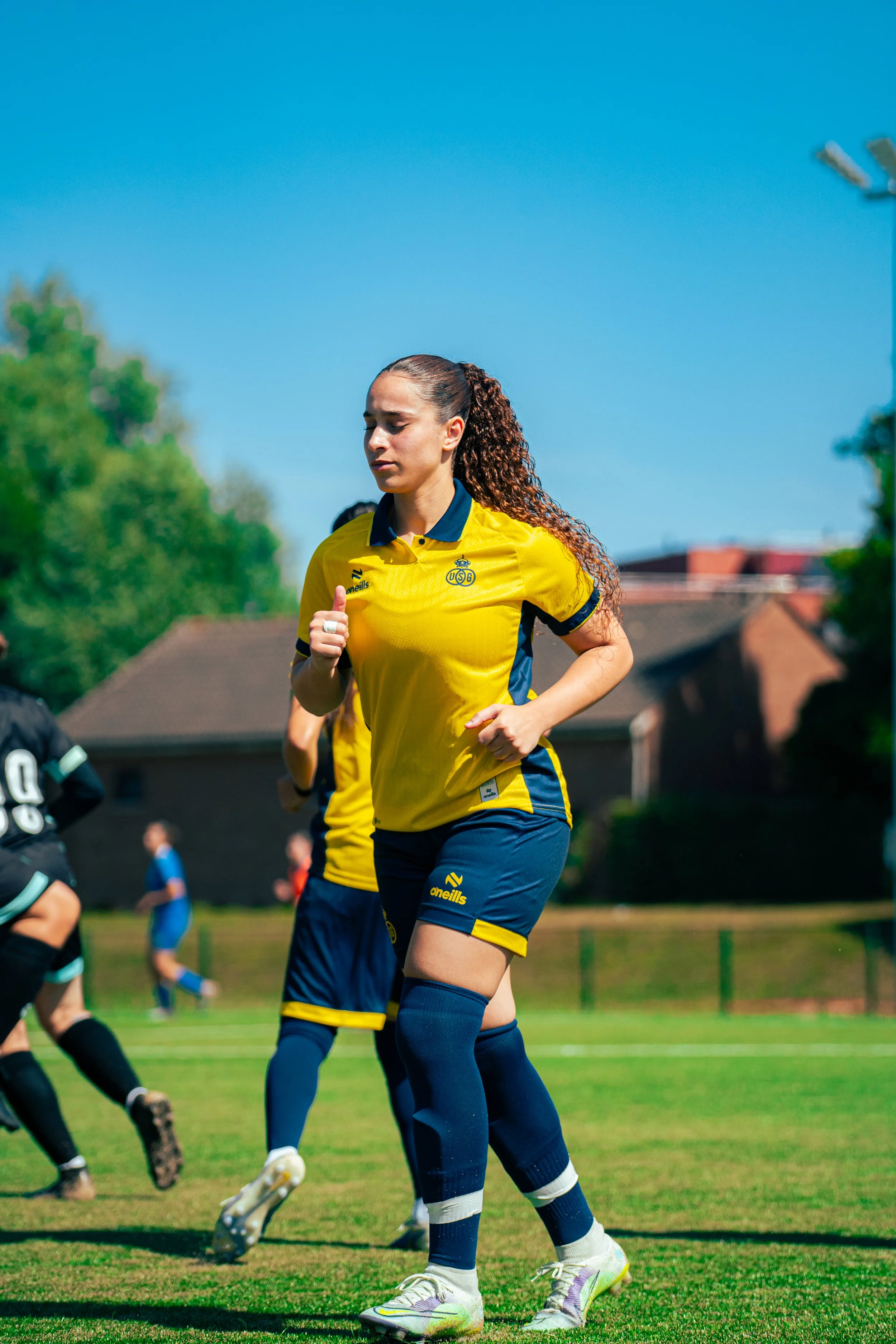 Female rugby player in yellow and blue uniform running on the field during a game under a clear blue sky, with other players and a building with trees in the background.