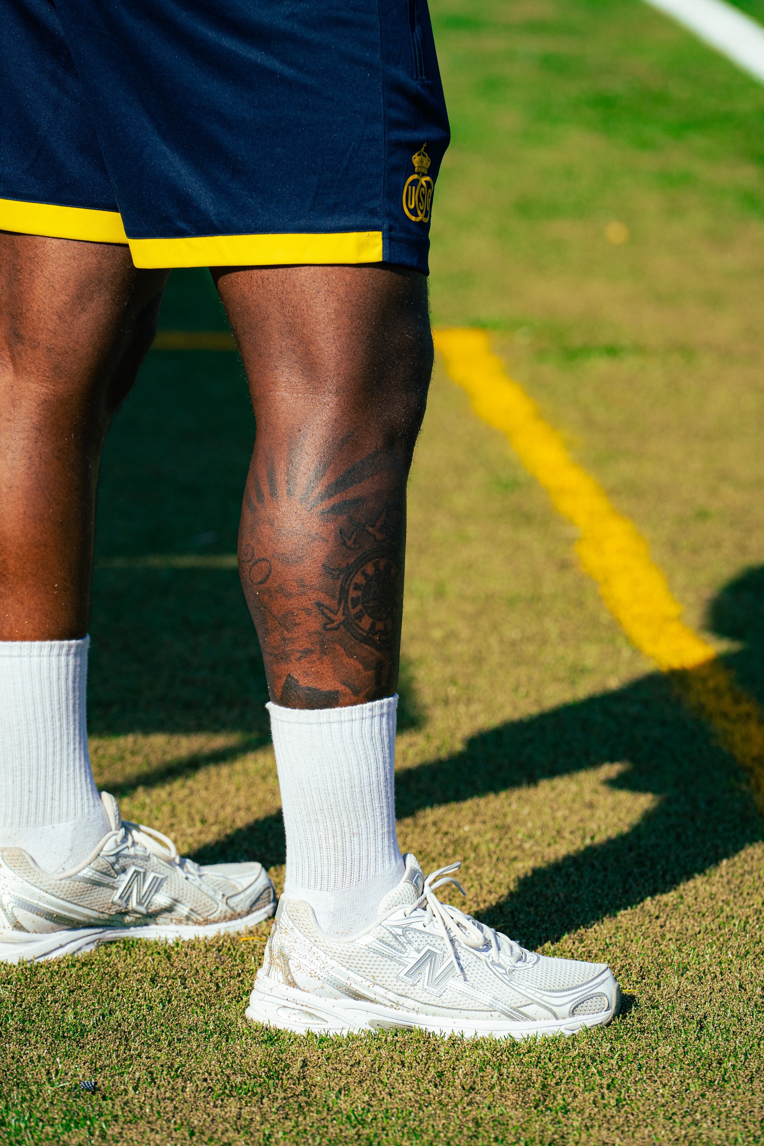 Close-up of a person's legs on a sports field, wearing white athletic shoes, white socks, navy shorts with yellow trim, and black athletic tattooed legs with a sun design tattoo on the right leg. The turf is green with a yellow line in the background