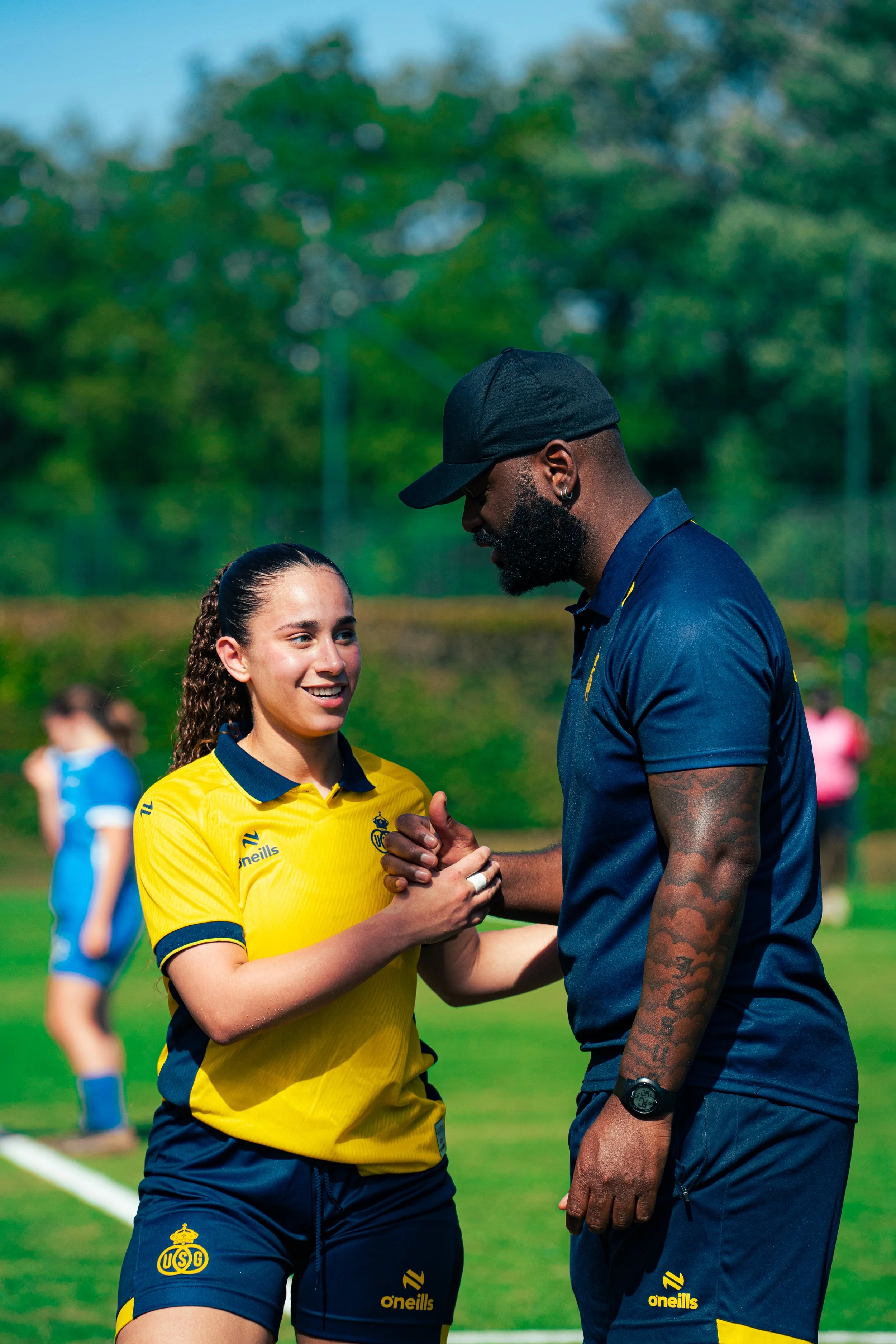 A female soccer player in a yellow jersey shaking hands with a male coach or teammate on a soccer field, with trees and other players in the background.