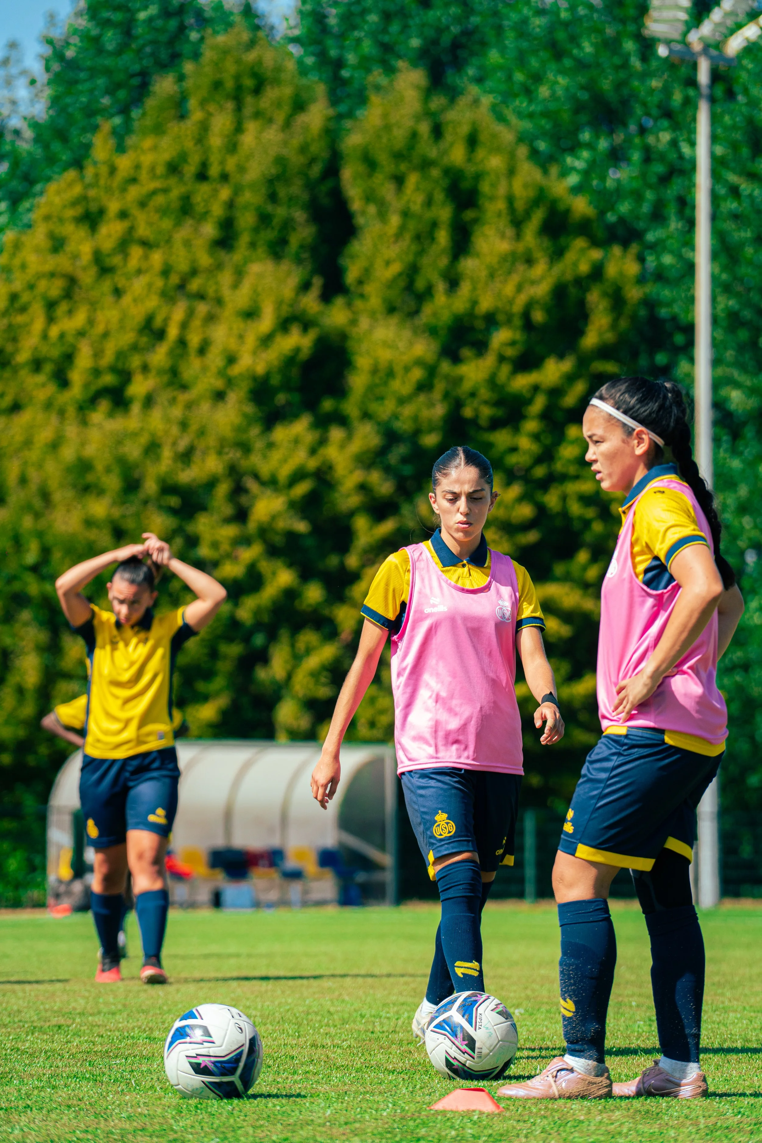 Women soccer players in yellow jerseys and pink training vests practicing on a grass field, with a soccer ball and orange cones, surrounded by trees.