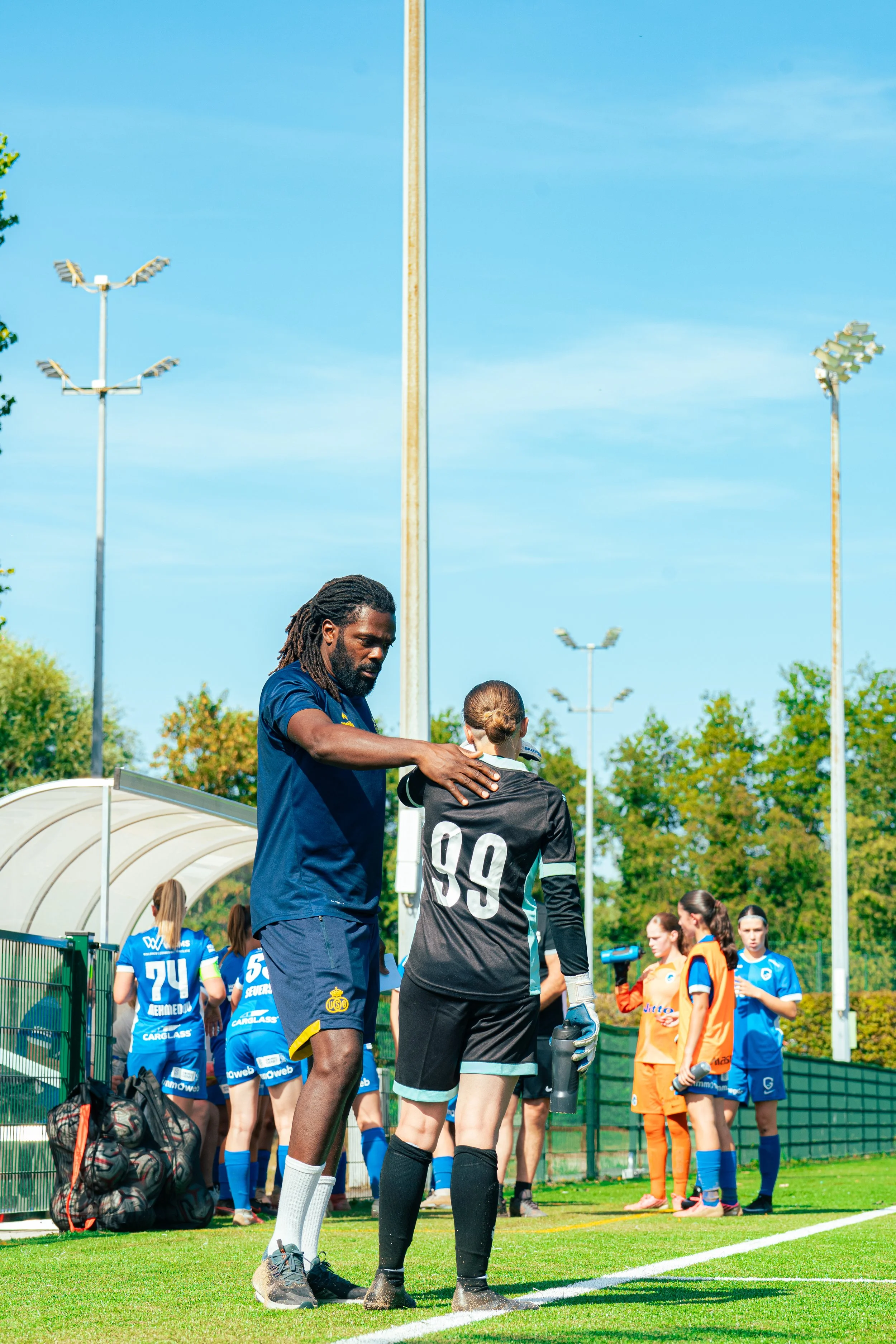 A soccer coach is talking to a goalkeeper with his hand on the goalkeeper's shoulder during a game or practice on a sunny day. Several players in colorful uniforms stand in line in the background near the soccer field, with trees and tall stadium lig