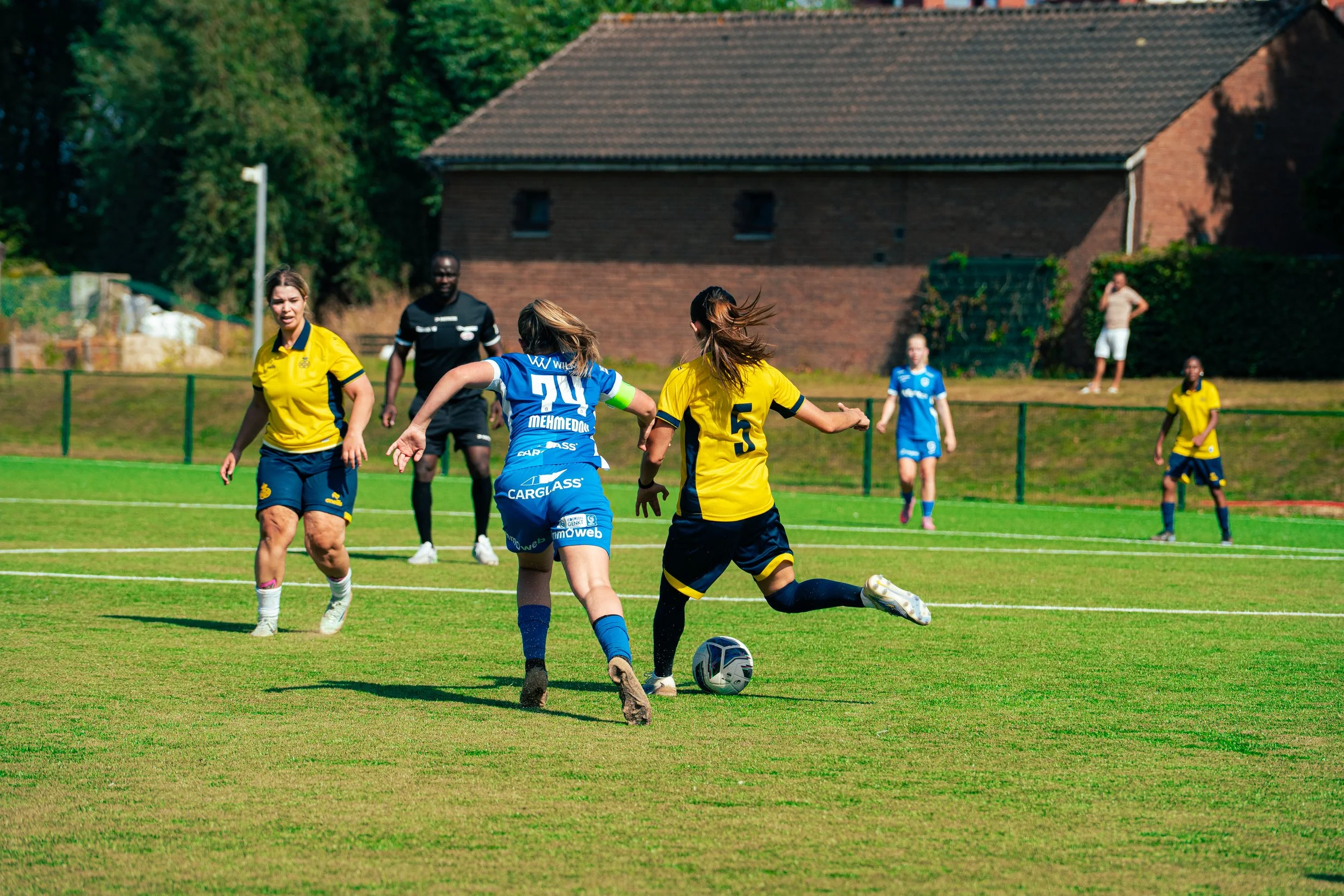 Women playing soccer on a grassy field during daytime. One woman in blue jersey with the number 20 and a captain's armband is in the center, about to kick the ball. Two women in yellow jerseys are nearby, one with the number 5, while other players an