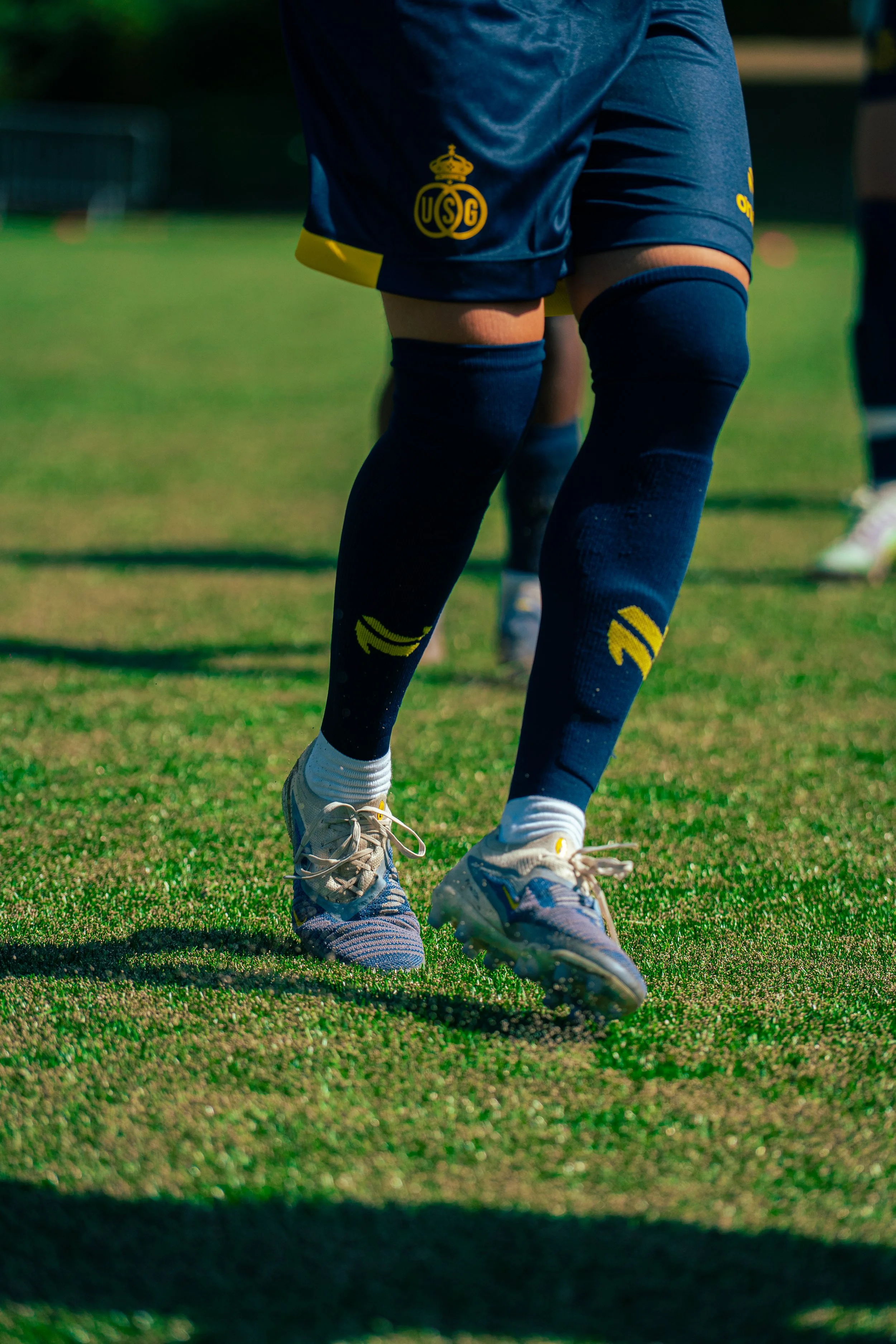 Close-up of a soccer player's legs wearing navy blue shorts with a yellow trim and the USG logo, navy blue knee-high socks with yellow design, and white athletic shoes on a grassy field.