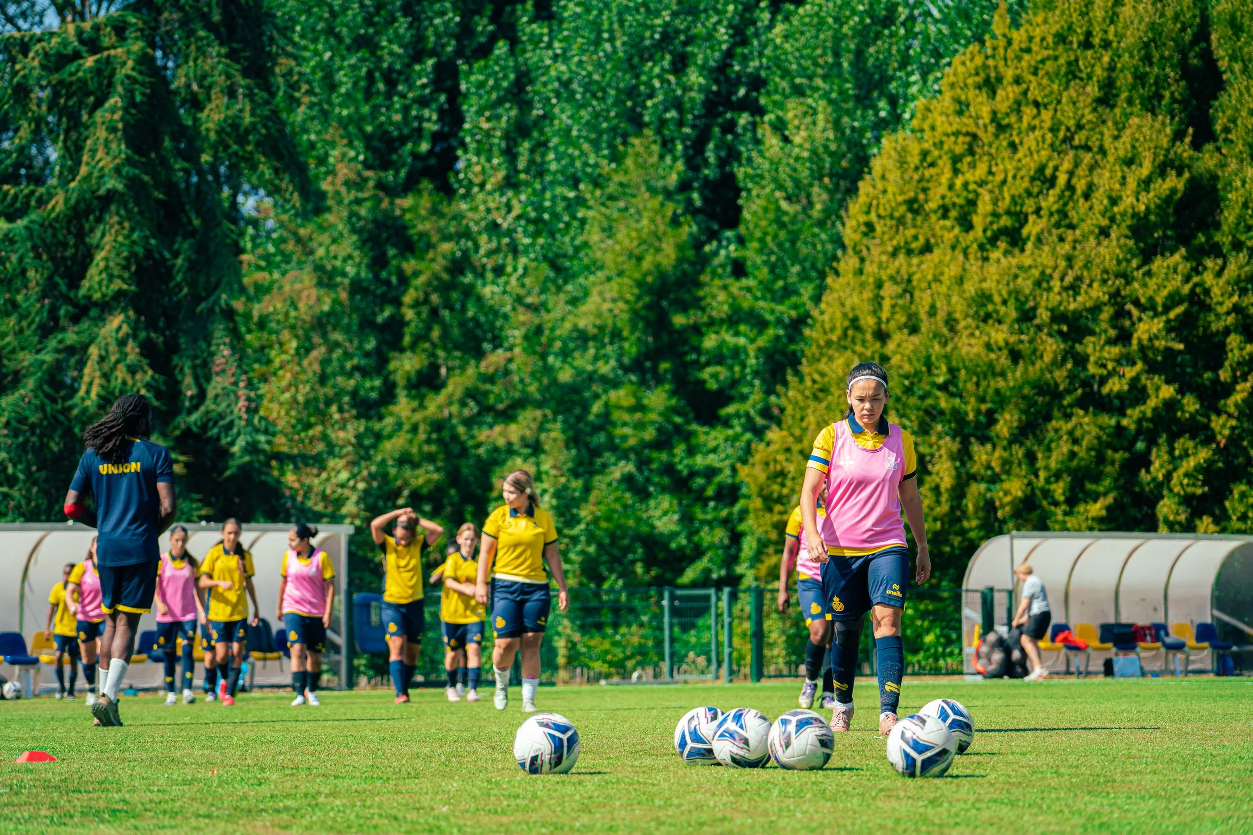 A women's soccer team warming up on a field, with several soccer balls in the foreground and players in yellow and pink training bibs preparing for practice under a green, wooded background.