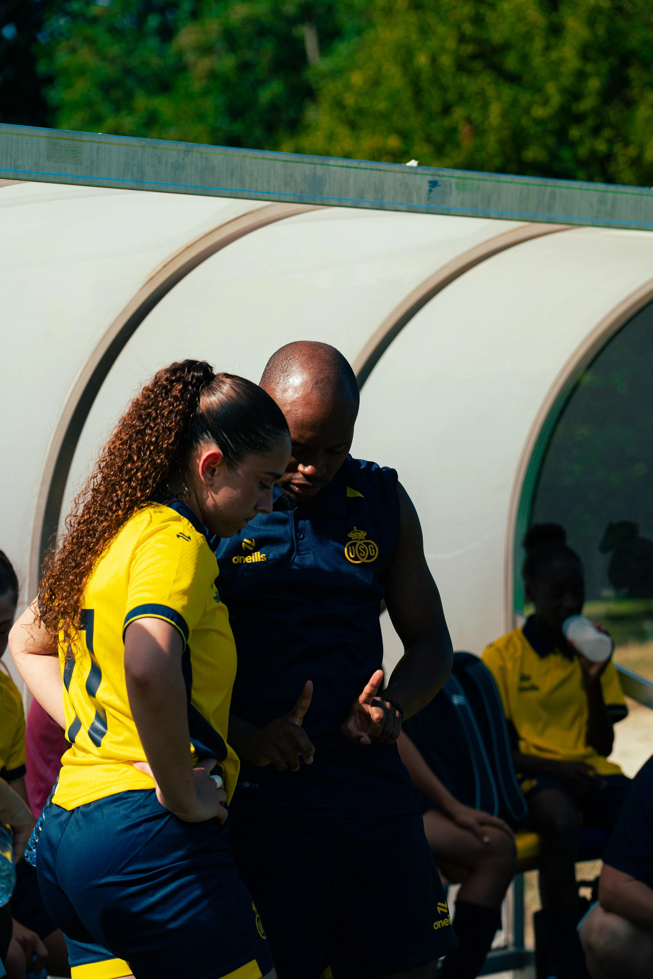 Two women stand outside on a bench, wearing sports uniforms, some with long hair, and are looking down, with one woman in a yellow shirt and one in a dark shirt with a crest. There are other people sitting on the bench and a girl with a disposable cu