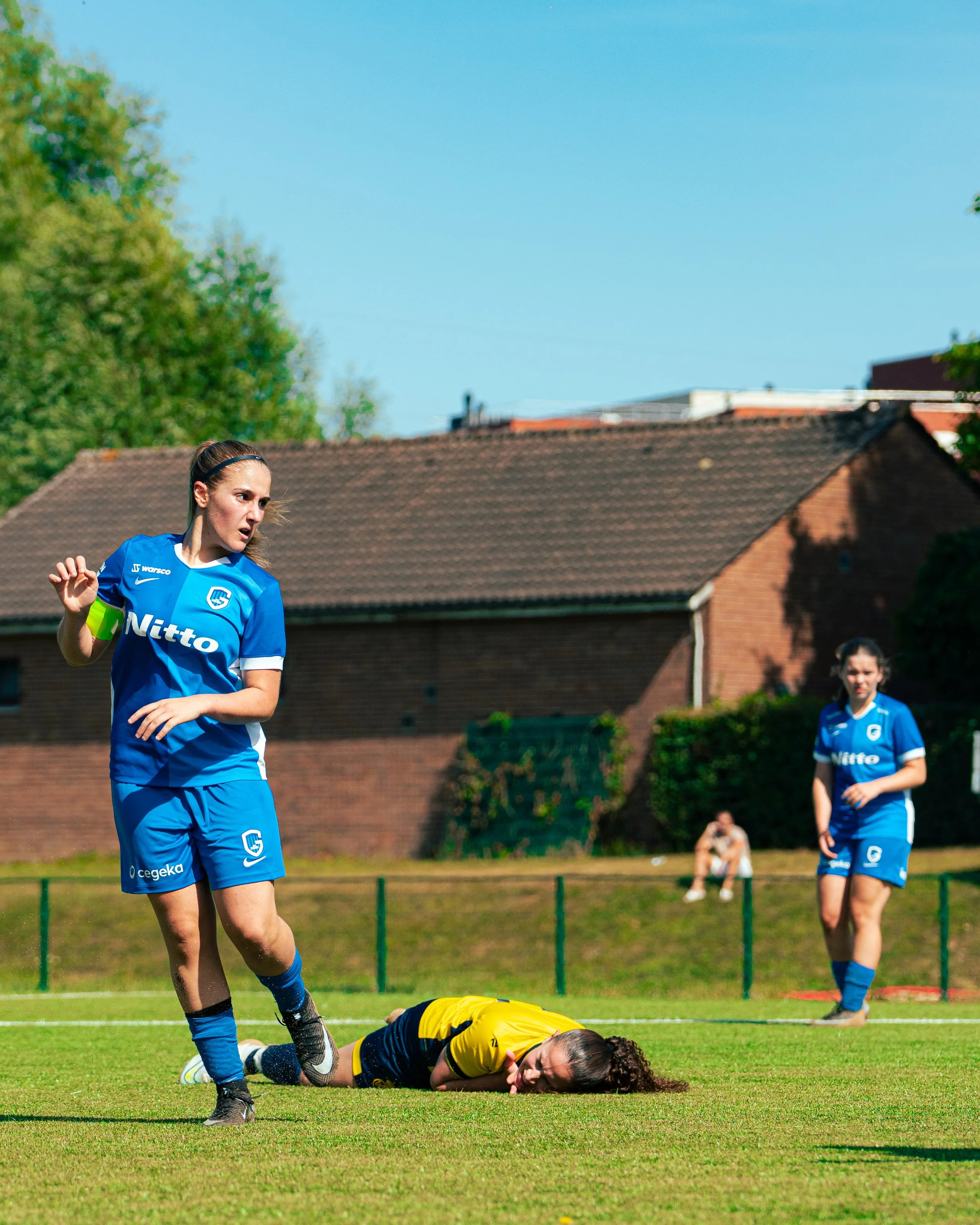 A female soccer player in a blue uniform is on the field, looking concerned after a tackle. A female goalkeeper in a yellow jersey is lying on the ground, clutching her knee, appearing injured. Two other players are in the background, one in a blue u