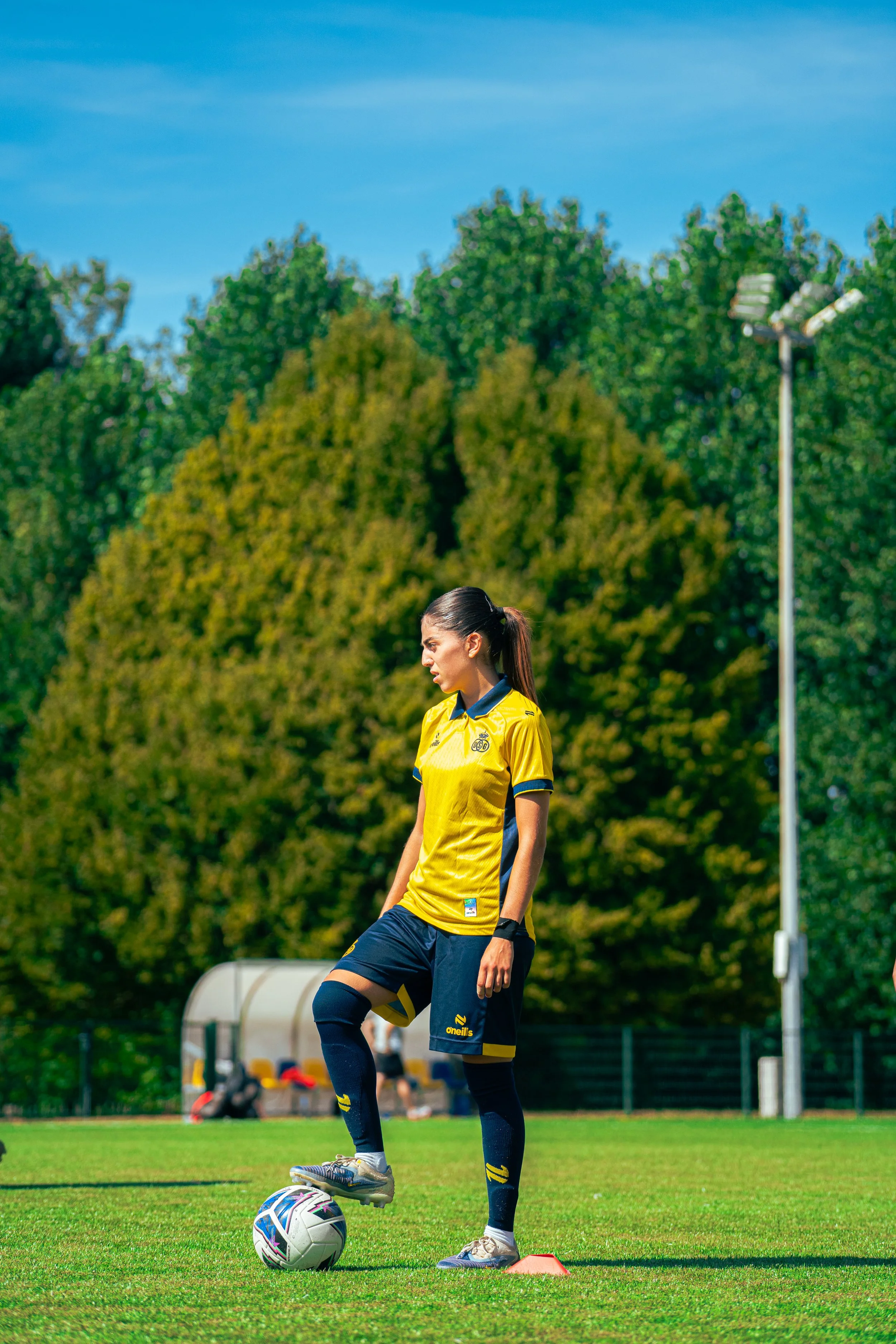 Female soccer player in a yellow jersey standing on a grassy field with a soccer ball at her foot, surrounded by trees and outdoor sports facilities.