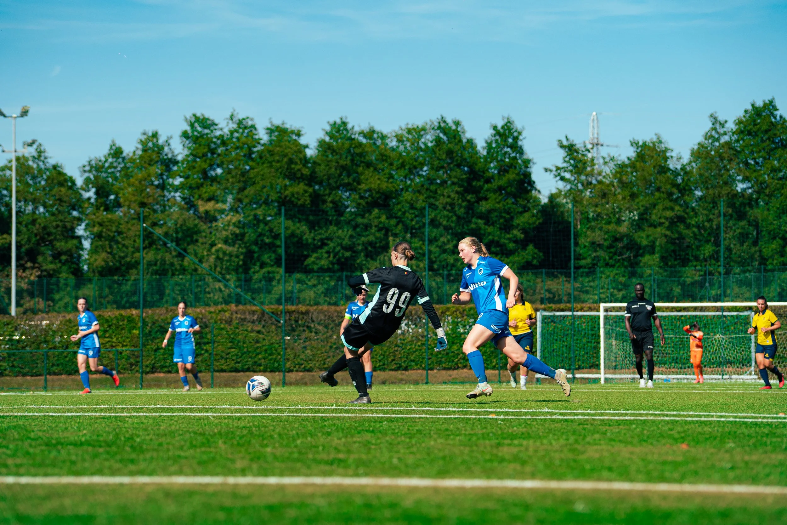 Women soccer players on a field, two players in black and blue jerseys competing for the ball, with other players and a goalkeeper in yellow in the background.