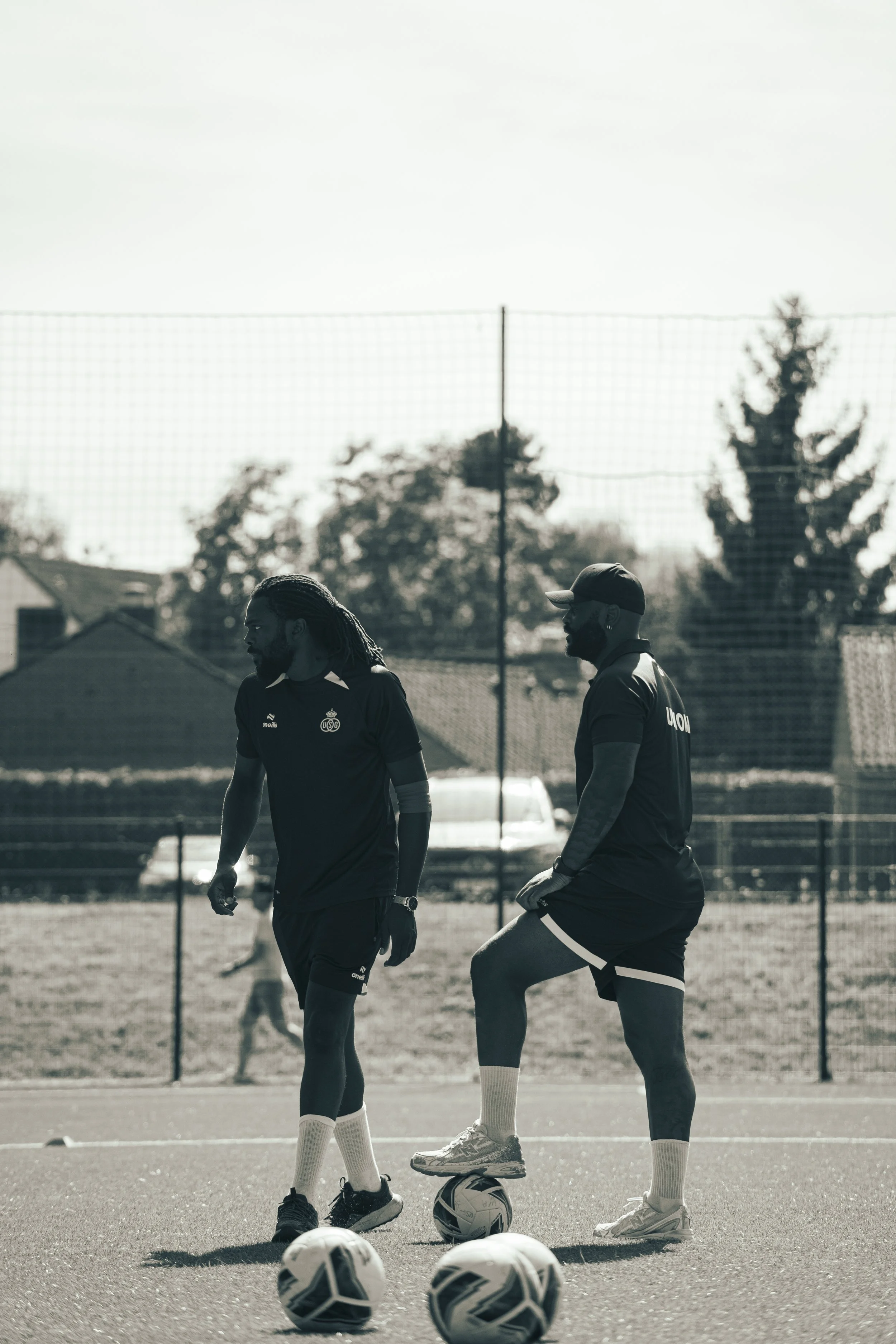 Two soccer players stand on a field with five soccer balls at their feet. One player has long dreadlocks and the other is wearing a cap. They are talking, with a fence and trees in the background.