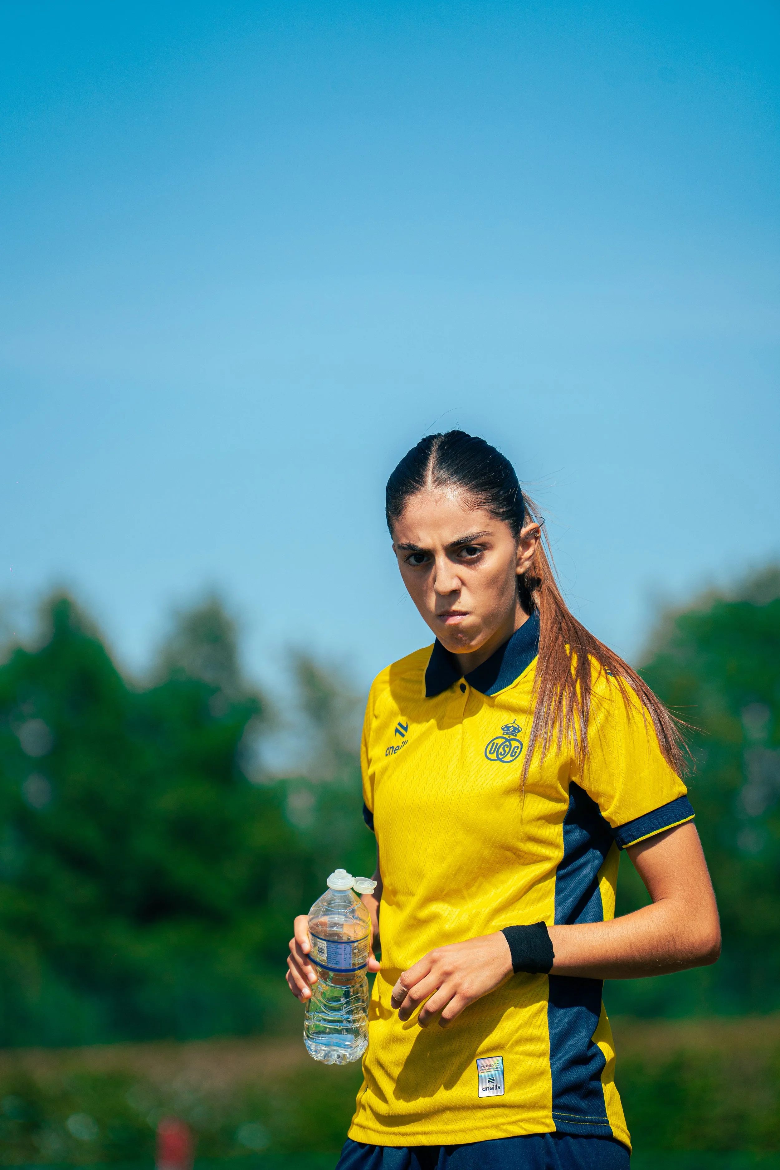 A female soccer player wearing a yellow jersey with a sports logo, holding a water bottle, standing outdoors with a blue sky and green trees in the background.