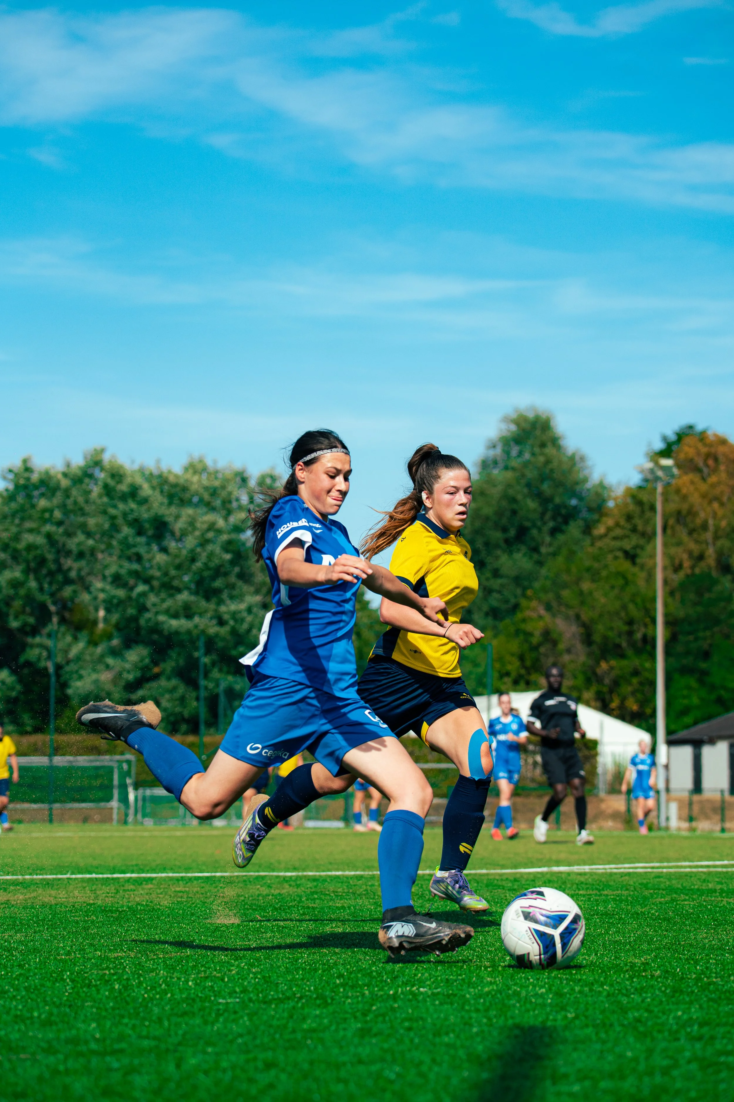 Two female soccer players, one in a blue uniform and the other in a yellow uniform, competing for the ball on a soccer field with trees and a blue sky in the background.