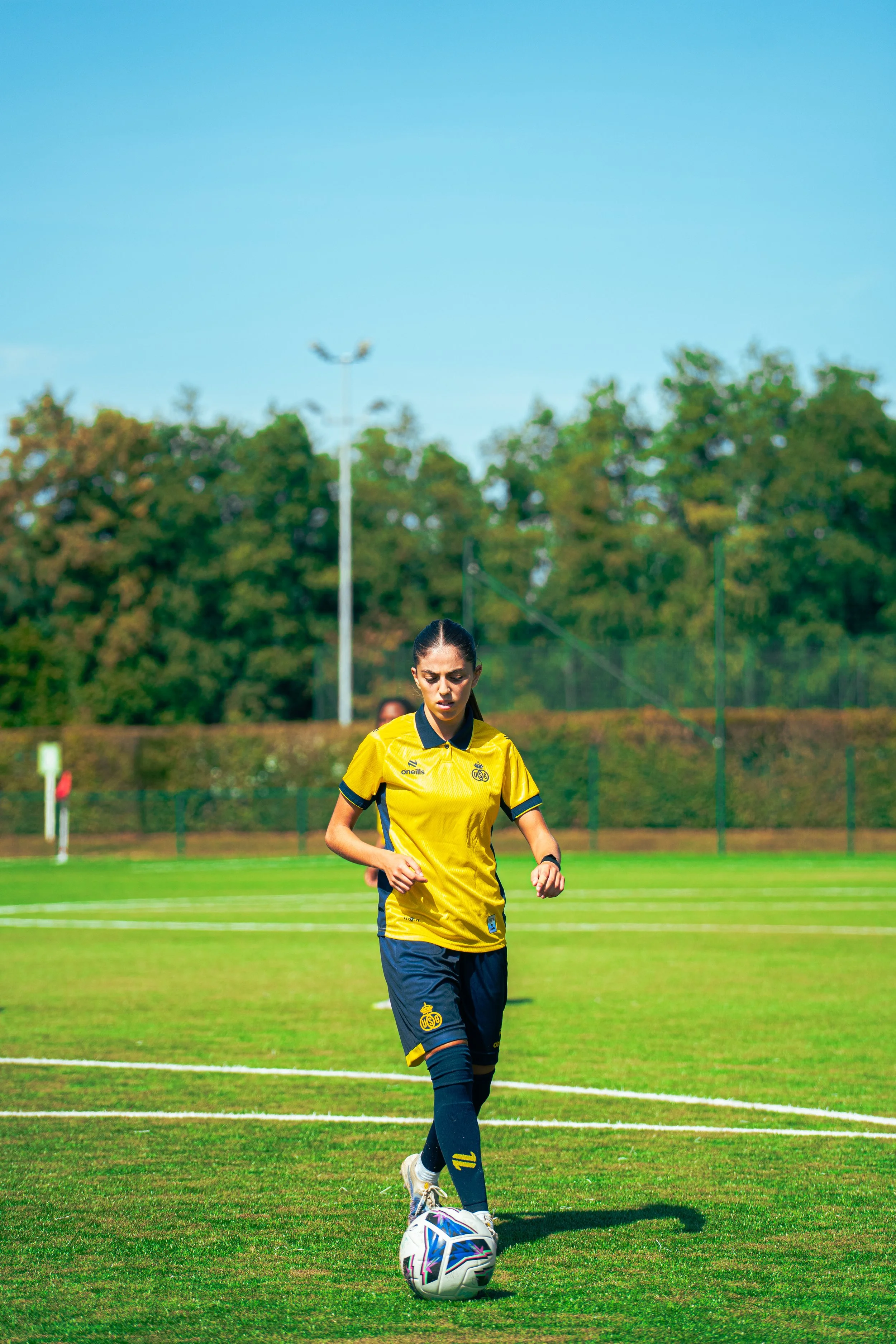 A female soccer player in a yellow and navy uniform is on a soccer field, preparing to kick a soccer ball.