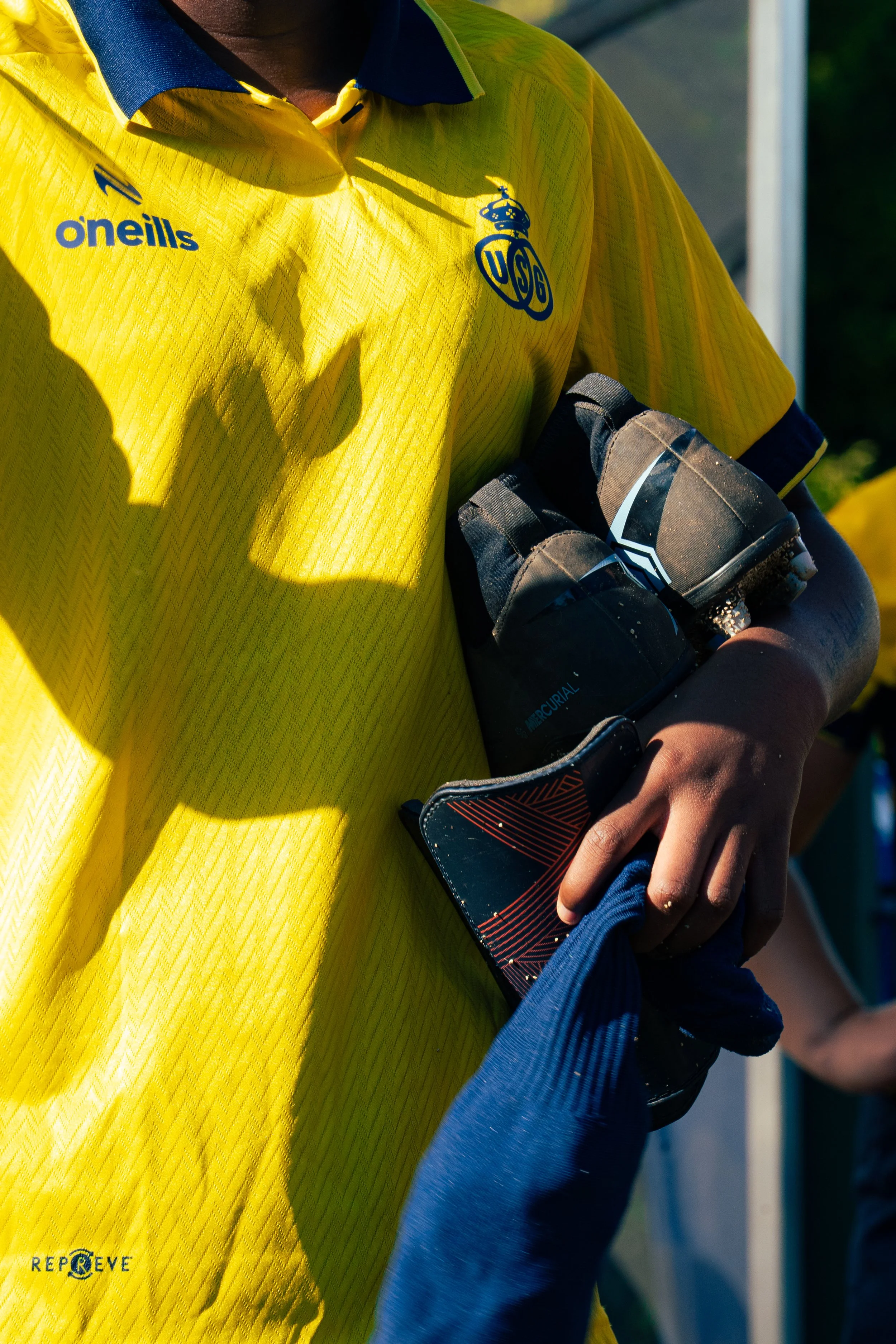 Close-up of a person in a yellow sports jersey holding a small water bottle and a navy blue item, with a blurred background.