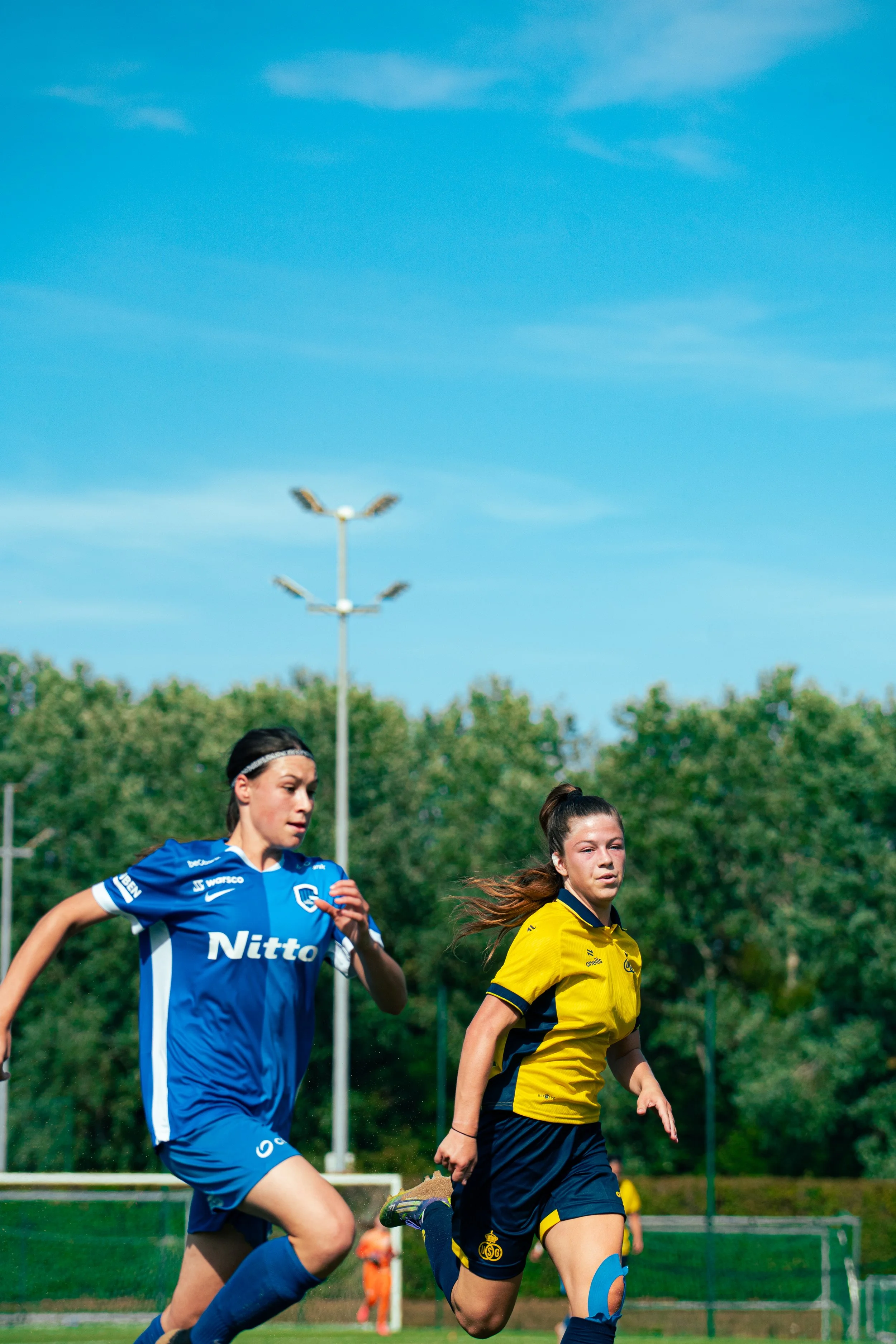 Two female soccer players running on the field during a match. One in a blue uniform and the other in a yellow uniform. The background features a green tree line, a clear blue sky, and stadium lights.