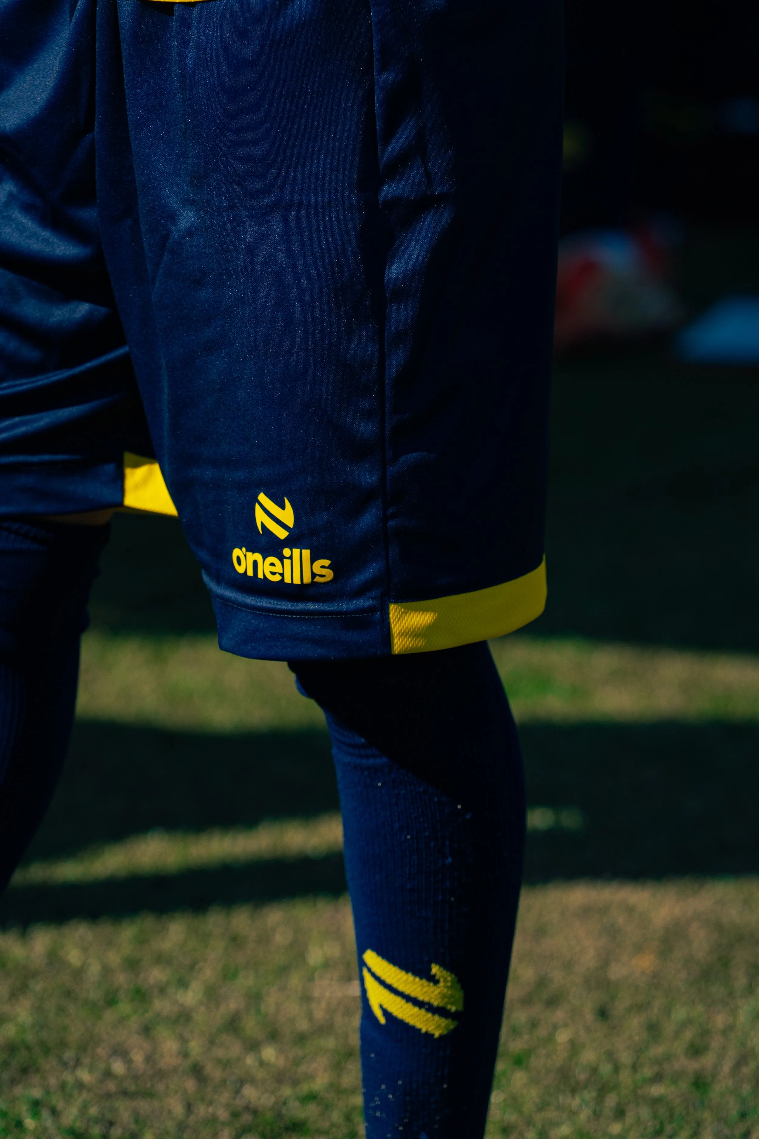 Close-up of a person wearing blue athletic shorts with yellow accents and the brand logo 'O'neills' on the side, black compression tights with yellow and black sports socks, standing on a grassy field.