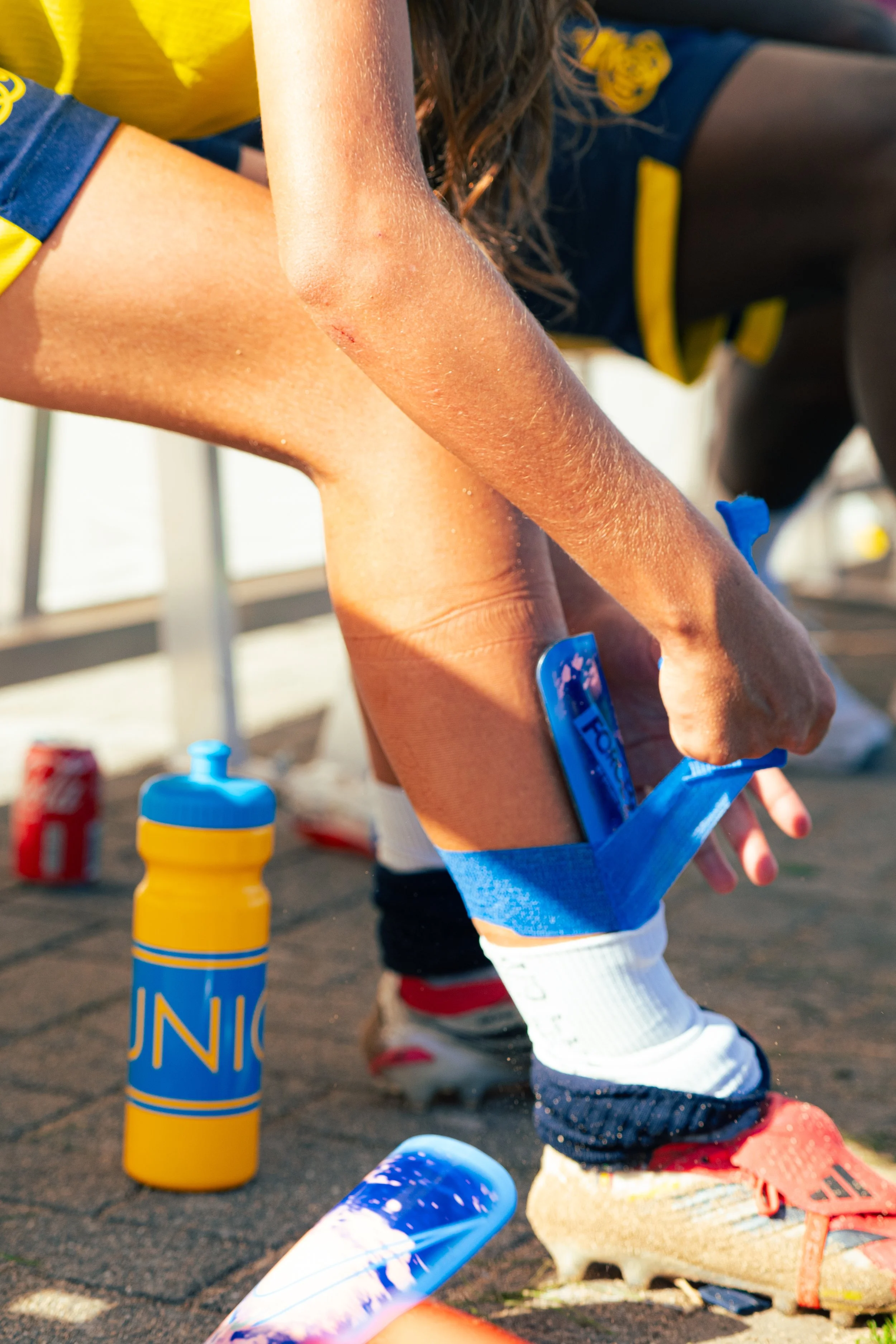 Close-up of a child's foot in a soccer sock and shoe, with a skinned knee visible on the arm. The scene appears to be at soccer practice or game, with sports water bottles and equipment visible on the ground.