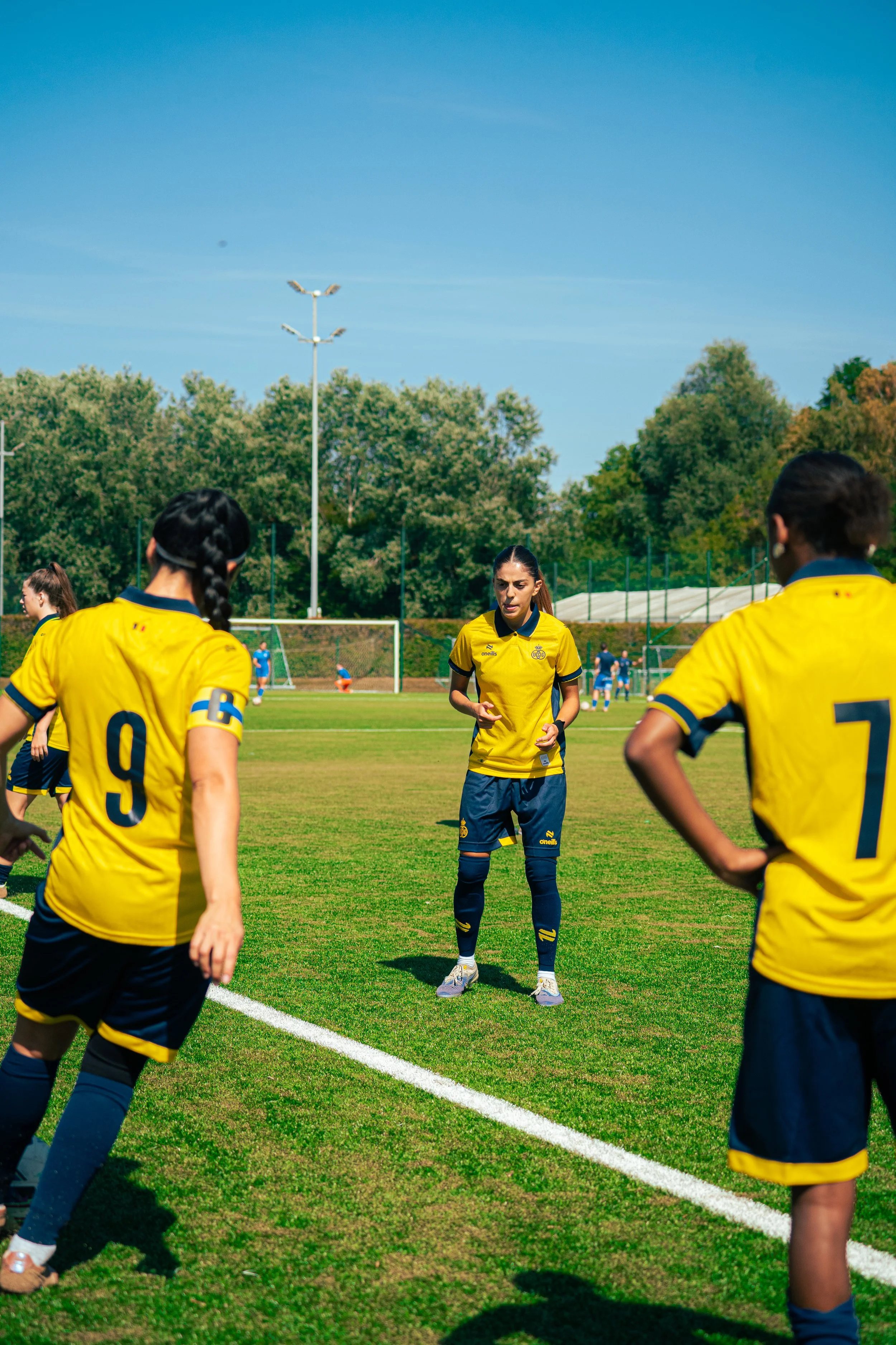 A group of female soccer players in yellow jerseys gathered on a soccer field during a game or practice. They are standing on green grass with a clear blue sky above and trees in the background.