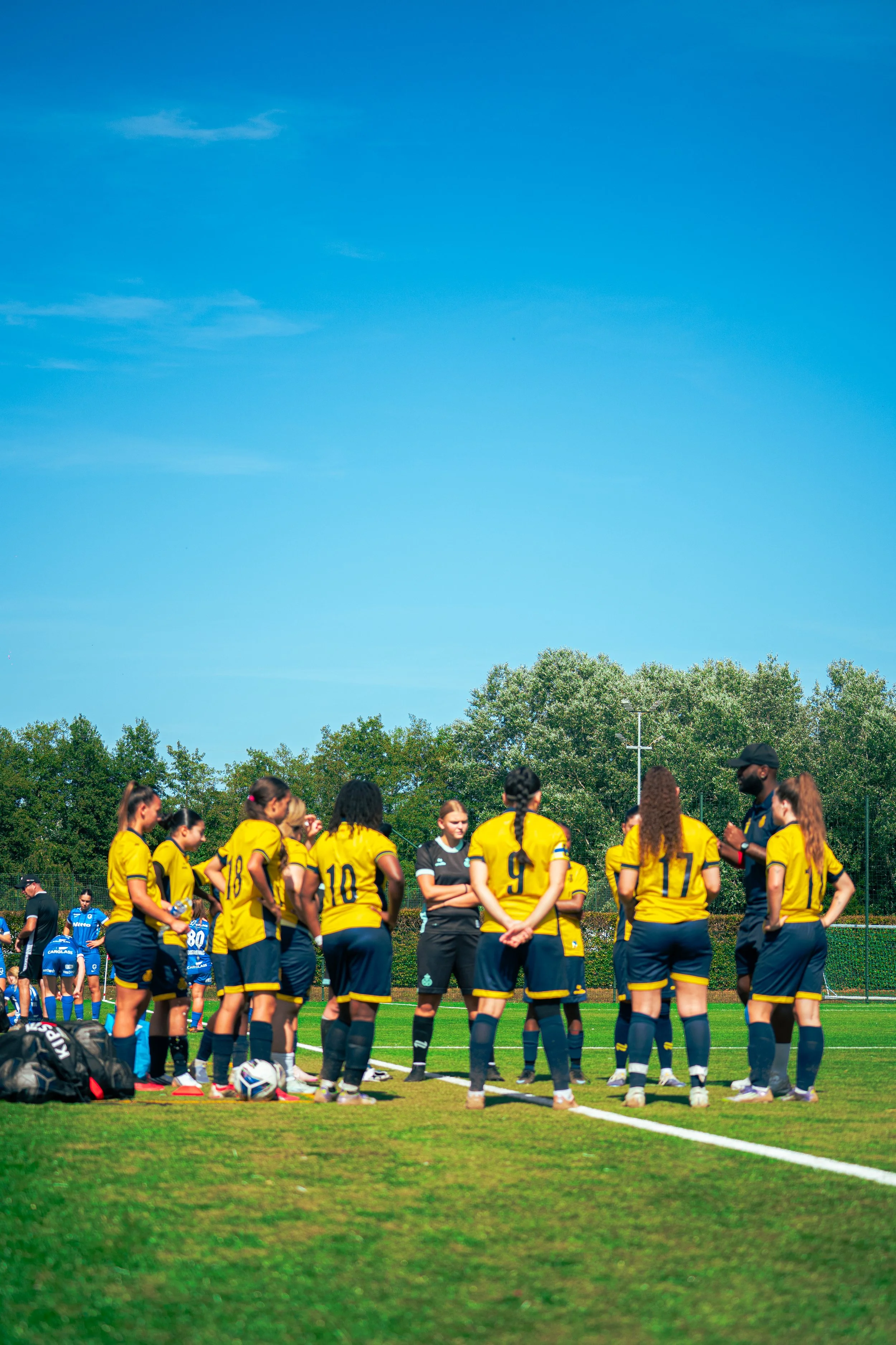 A group of female soccer players wearing yellow jerseys and black shorts are standing in a huddle on a soccer field during daytime. People are visible in the background, with trees and a clear blue sky overhead.