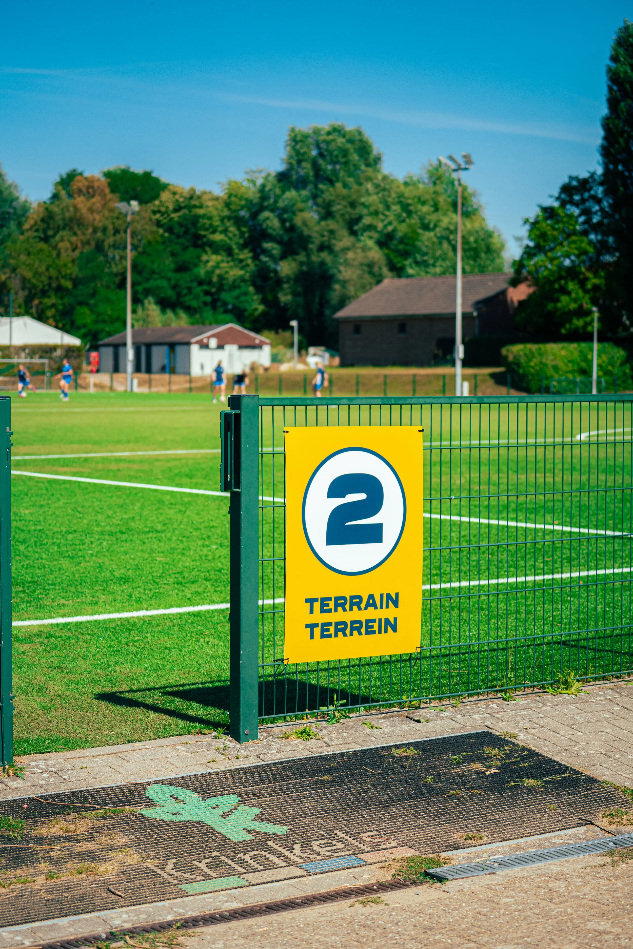 Soccer field with players and a yellow sign on the gate that reads 'Terrain Terrein', with trees and small buildings in the background.