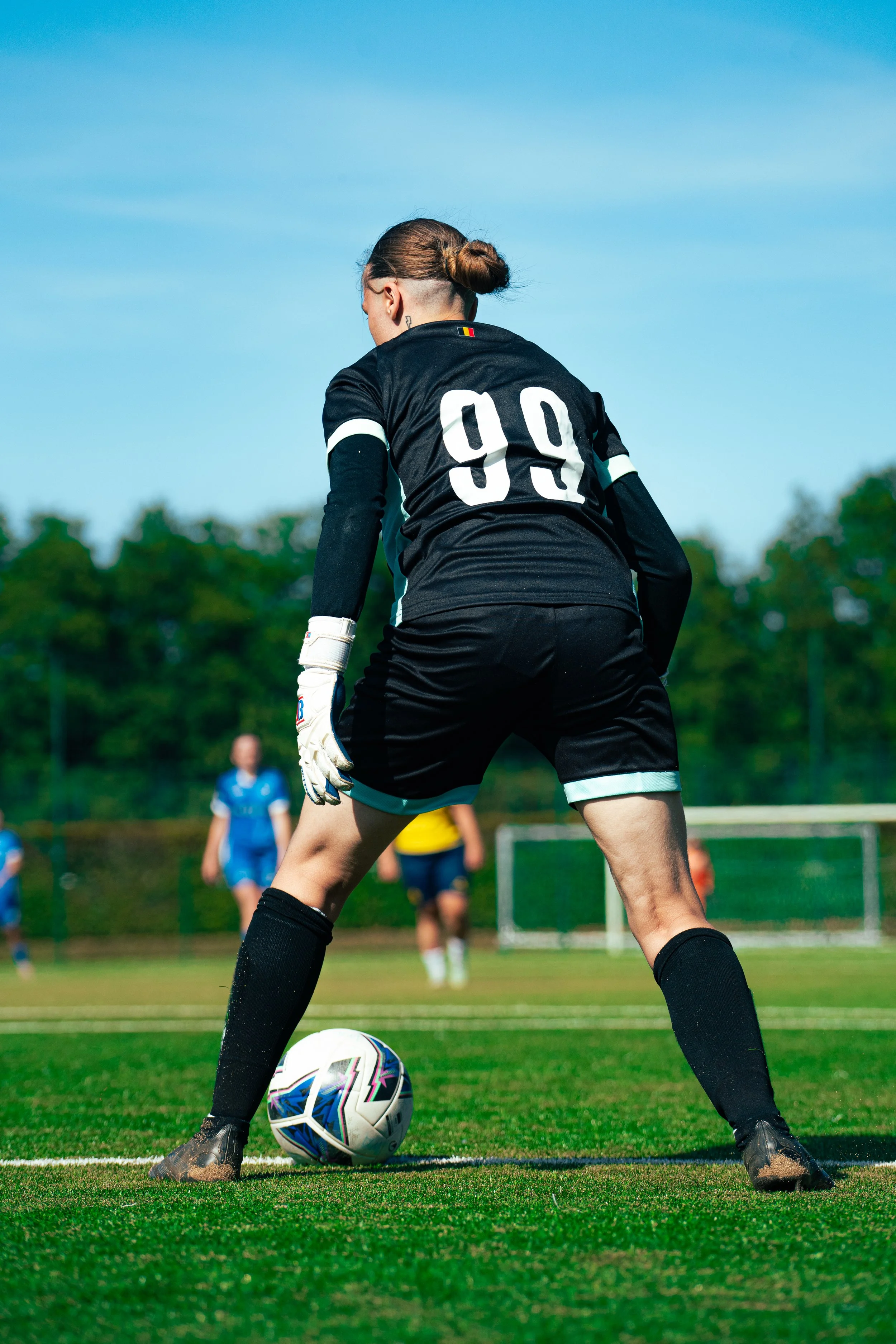 A female soccer goalkeeper in black jersey with the number 99, wearing gloves, preparing to kick a ball on a green field during daytime, with other players and goals visible in the background.