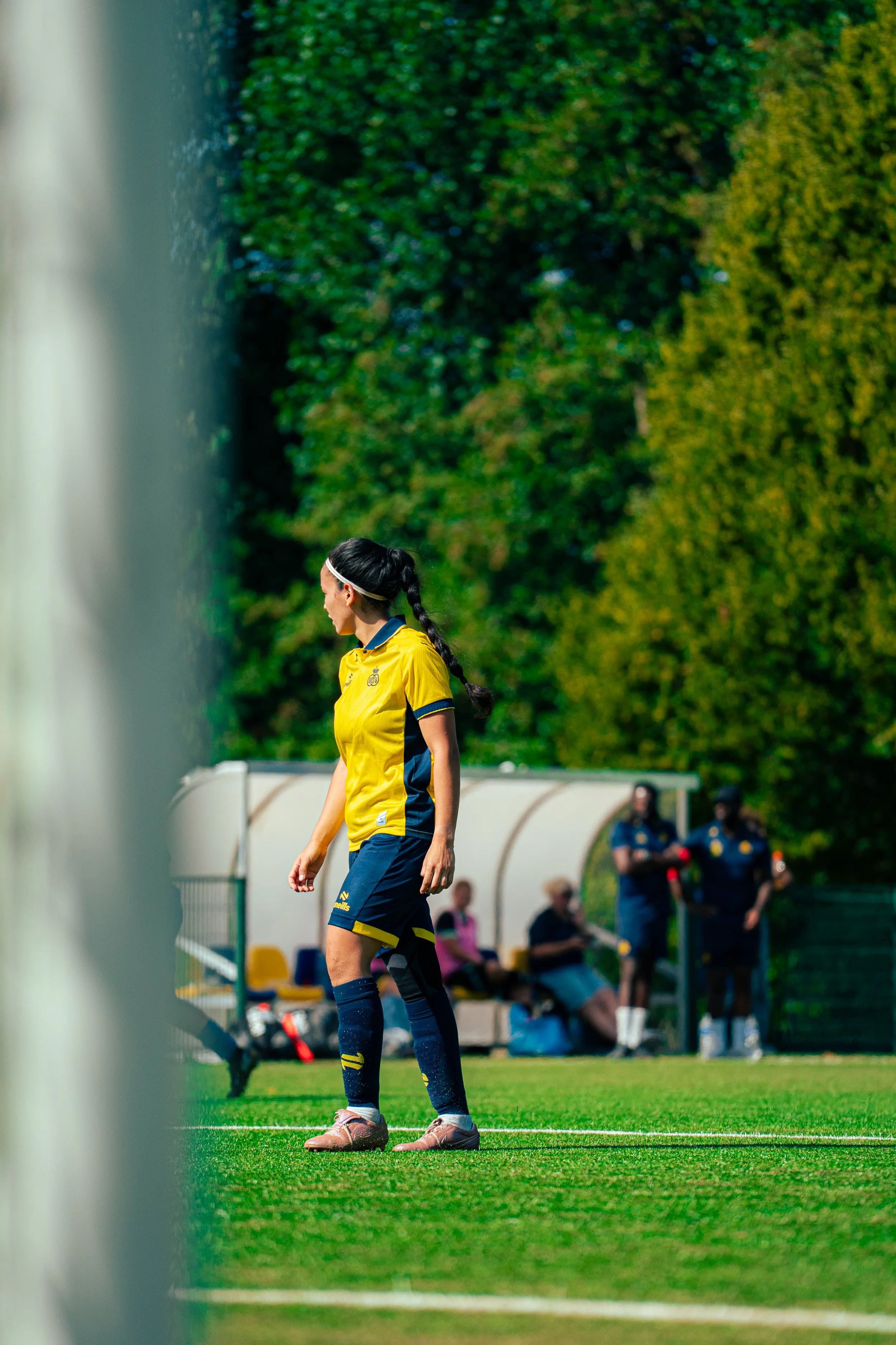 A female soccer player in yellow and blue uniform standing on the sidelines of a soccer field, with a green background of trees. She is wearing a headband, and her hair is in a braid. Several people are sitting on a bench in the background, with othe