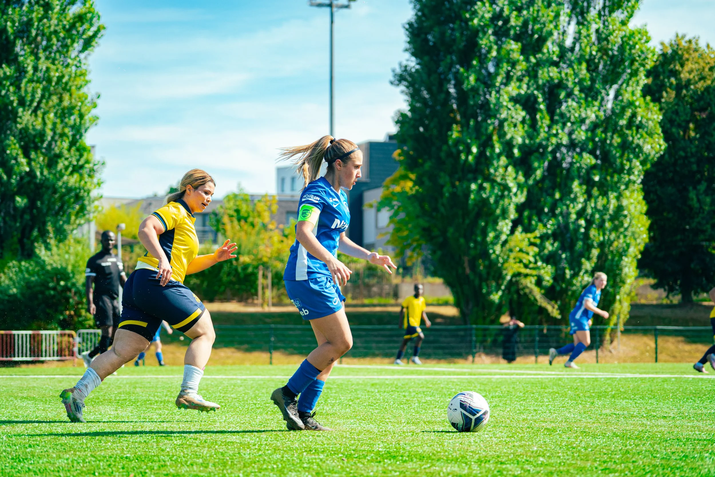 Women playing soccer on a green field with trees in the background during daytime.