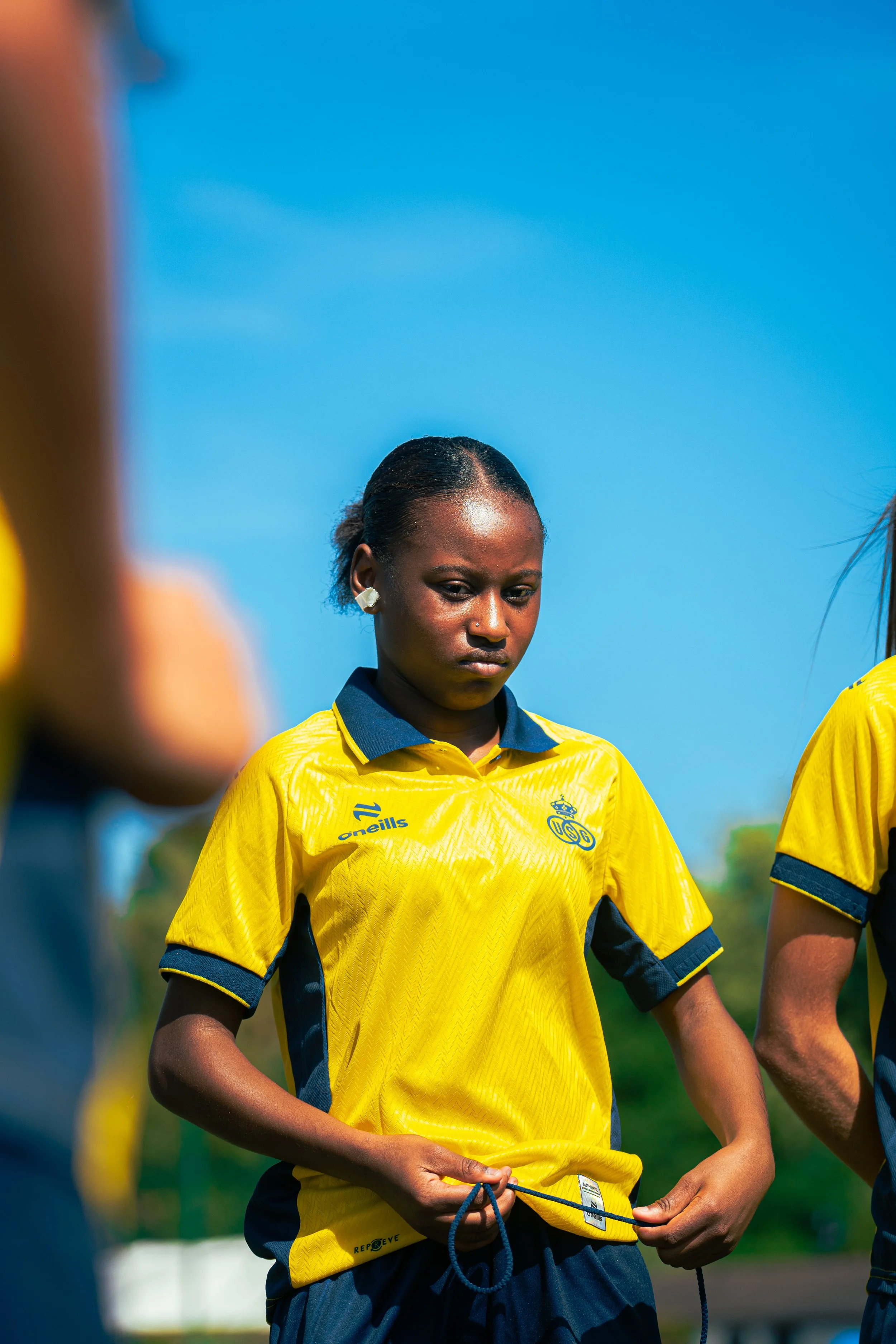 A young woman wearing a yellow sports jersey with black and yellow accents, holding a blue rope in her hands. She has earrings and a headset, with a serious expression, and there are blurred figures in yellow jerseys on either side of her against a b