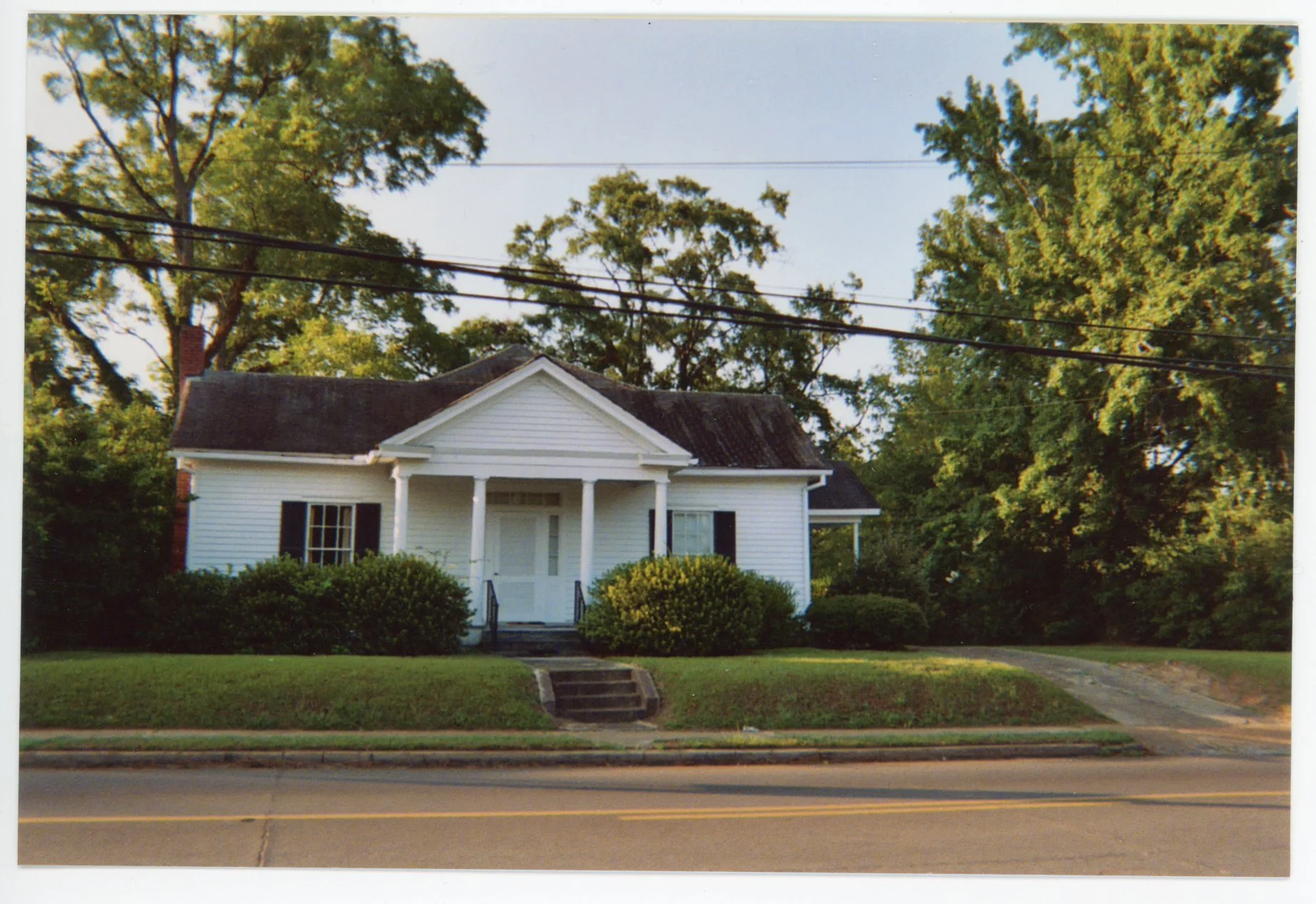 A white house with a front porch, black shutters, and a chimney, surrounded by green bushes and trees, with a clear blue sky in the background, along a street with a sidewalk and power lines overhead.