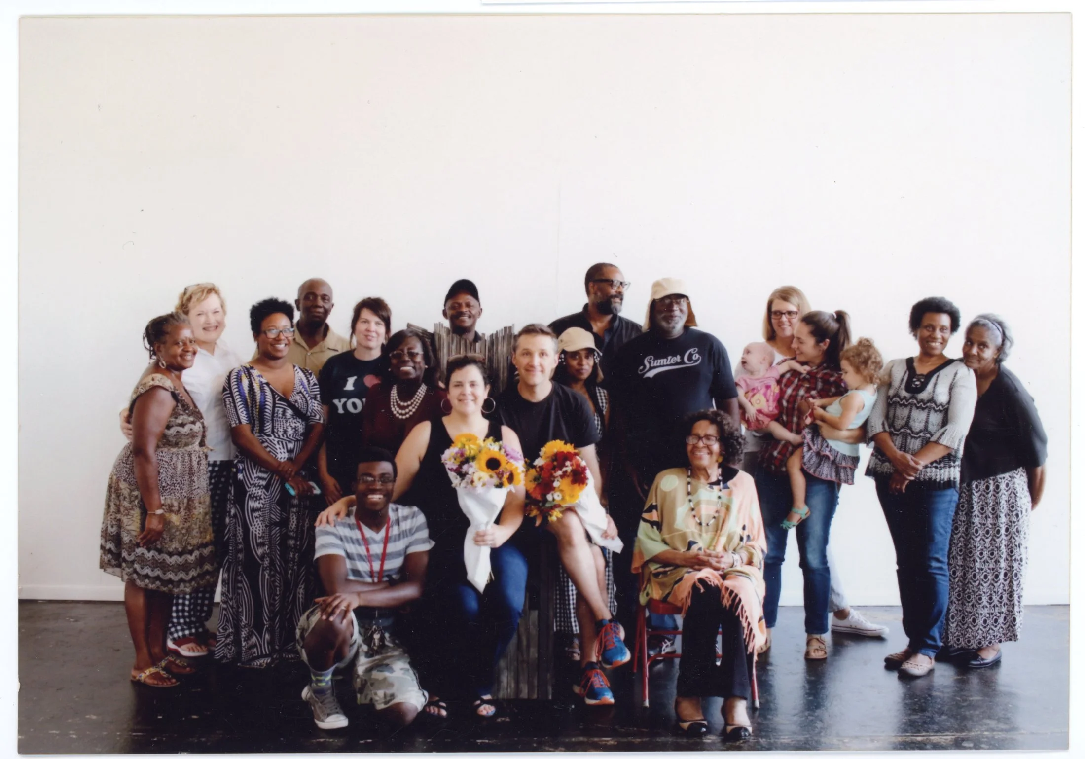 Group of diverse people standing together indoors against a plain white wall, some smiling, with a woman in front holding a bouquet of flowers.