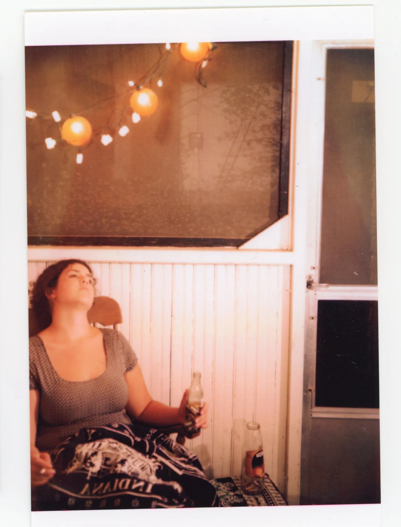 A woman with short dark hair reclining on a chair, holding a bottle in her right hand. She is wearing a patterned top and pants, with her eyes closed. The setting appears to be a porch or outdoor area with white wooden paneling and a window, under wa