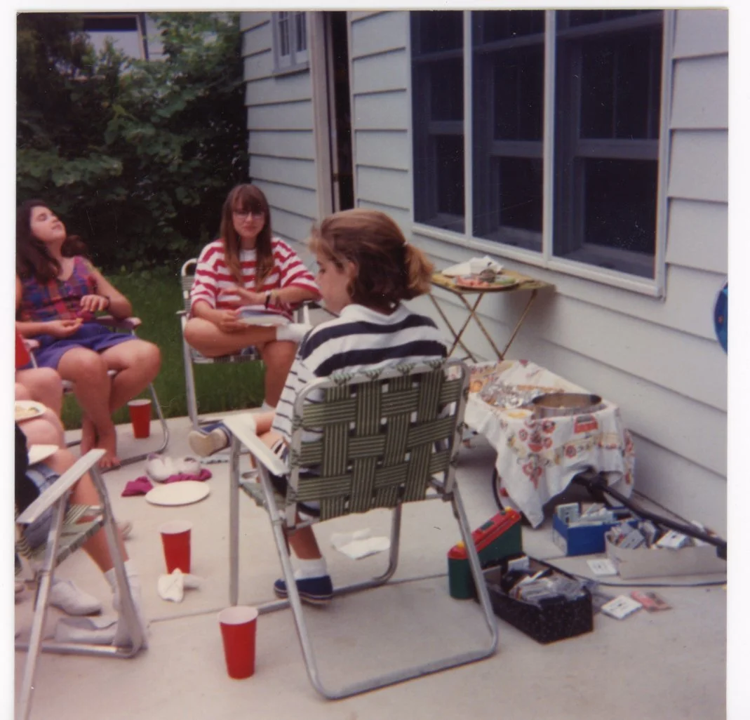 A group of children sitting outside on a patio, with one girl facing away, wearing a striped shirt, and others sitting on chairs or the ground, with red cups and snacks around, near a house with blue siding and open windows.