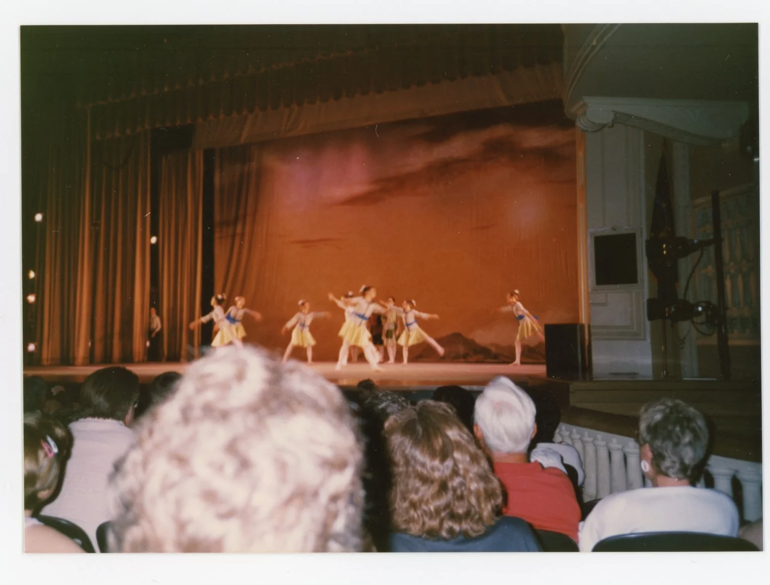 A group of dancers performing on stage with a scenic backdrop and audience members watching.