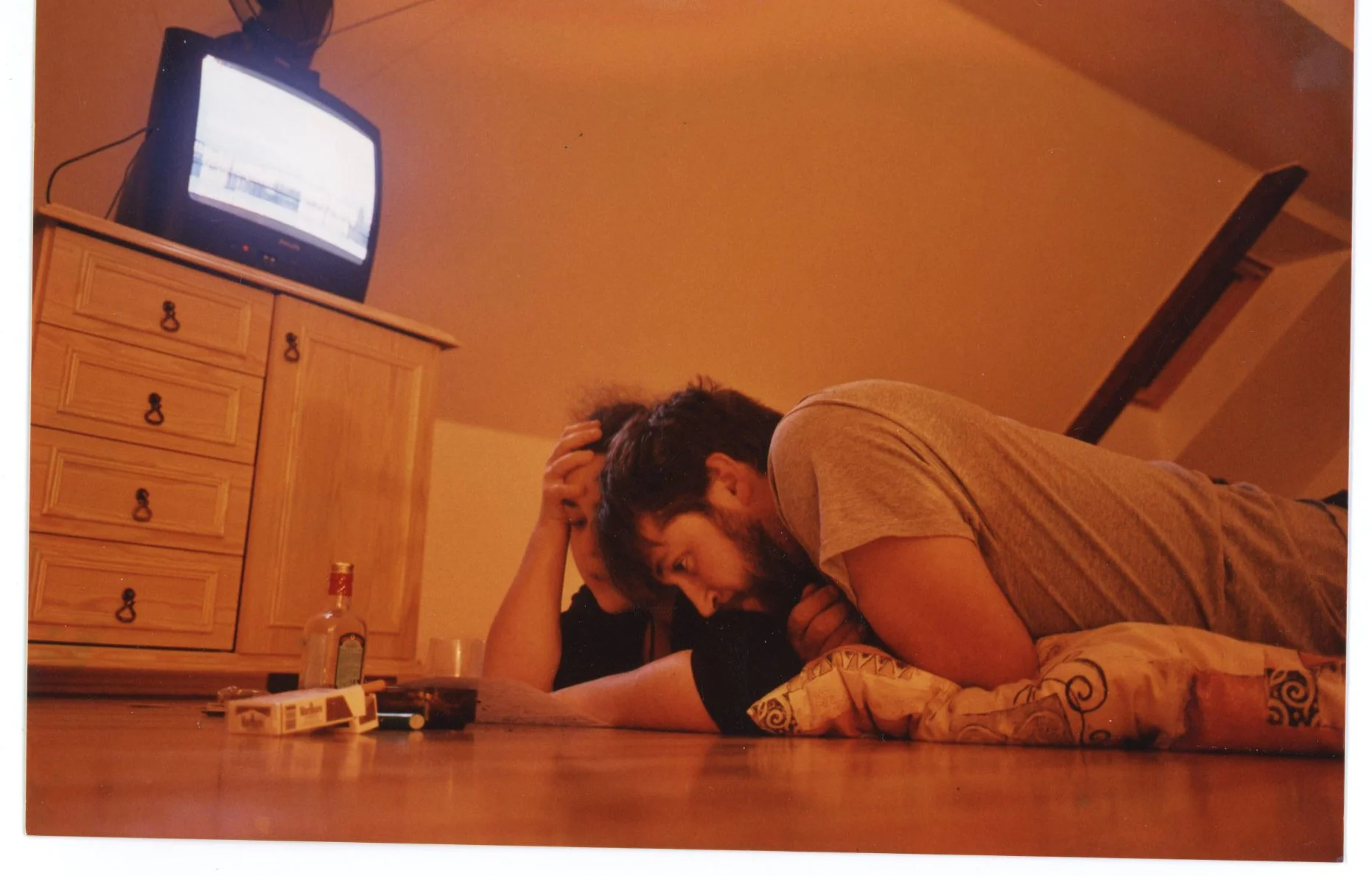 A man with a beard lying on the floor, resting his head on his hand, with a woman lying beside him, resting her head on his arm in a room with a wooden dresser and an old television.
