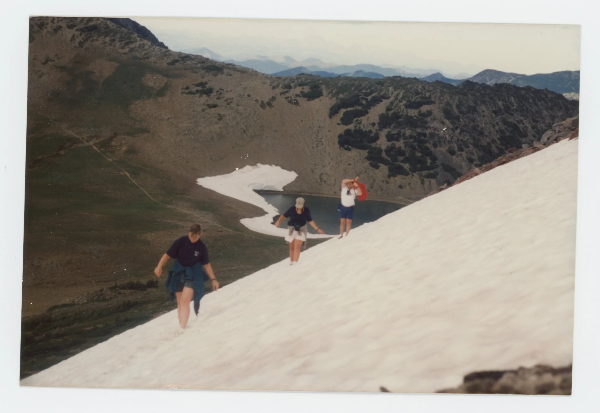 People hiking on a snow-covered slope in a mountainous area with rocky peaks and sparse vegetation.