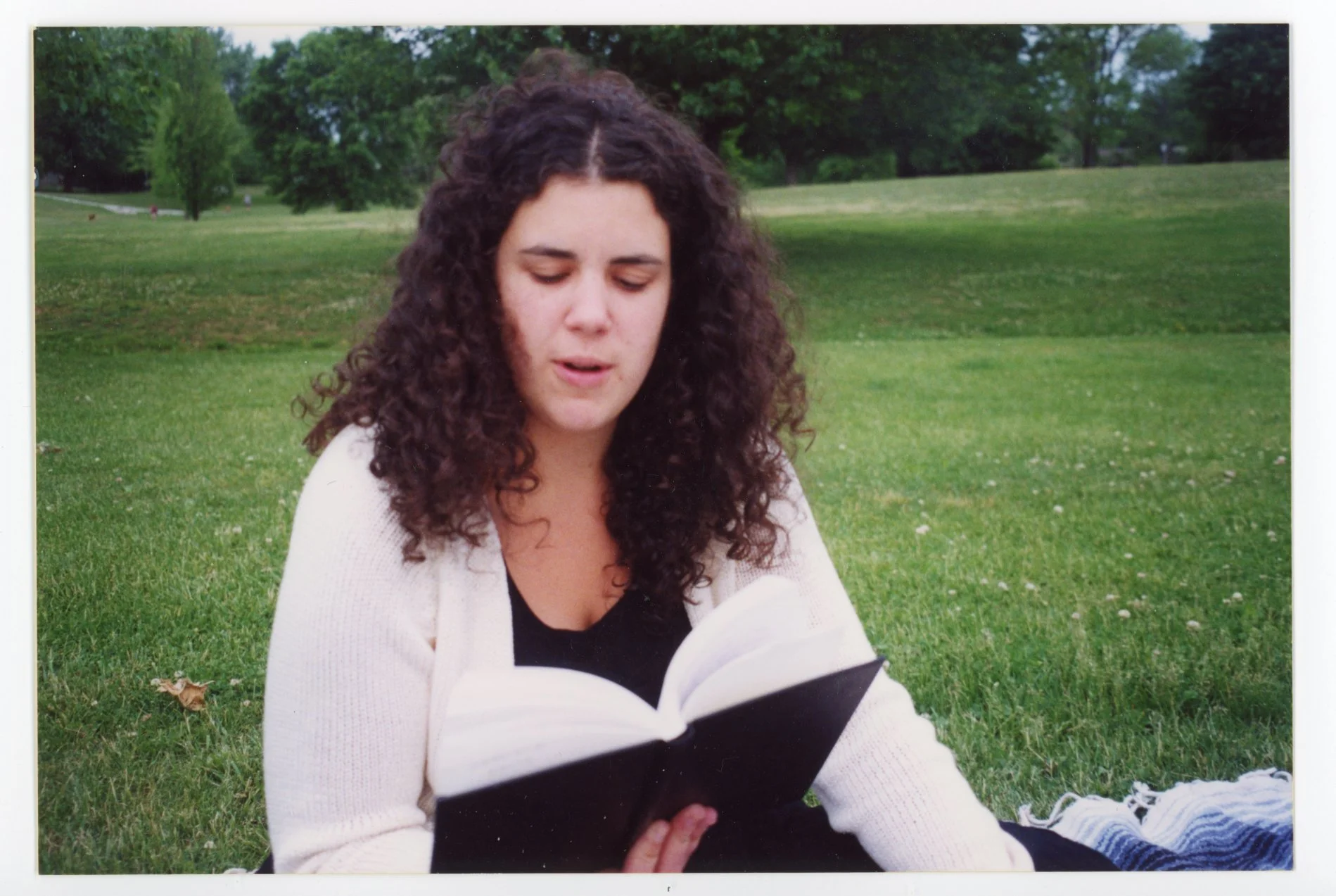 Young woman with dark curly hair sitting on grass in park reading a book, wearing a black top and white cardigan.