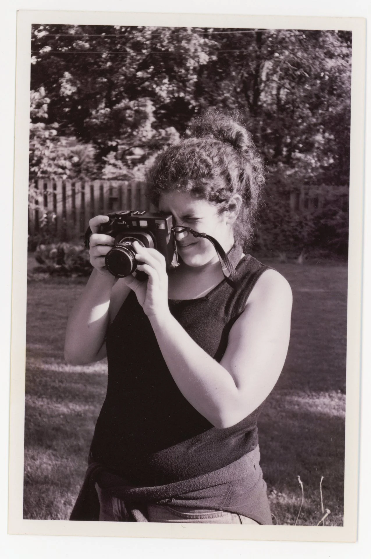 A black and white photo of a woman with curly hair taking a photograph outdoors with trees and a wooden fence in the background.
