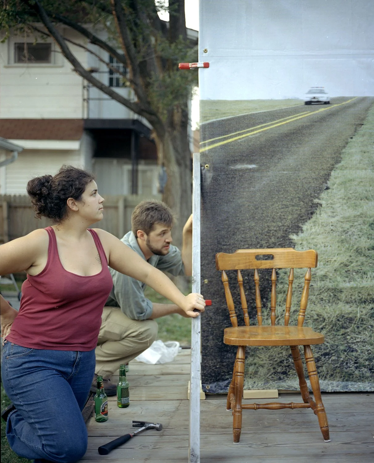 Two people, a woman and a man, appear to be setting up a photo backdrop with an outdoor scene of a road and grass. The woman is wearing a red tank top and blue jeans, and the man is wearing a gray shirt and beige pants. They are kneeling on a wooden 