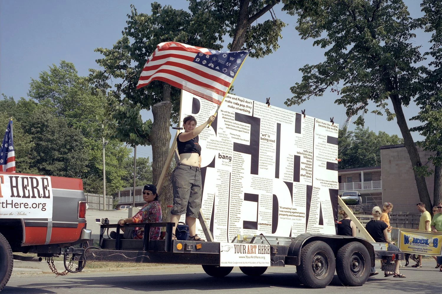 Protest parade float with large art installation, American flags, and a woman holding a flag, surrounded by people and trees.