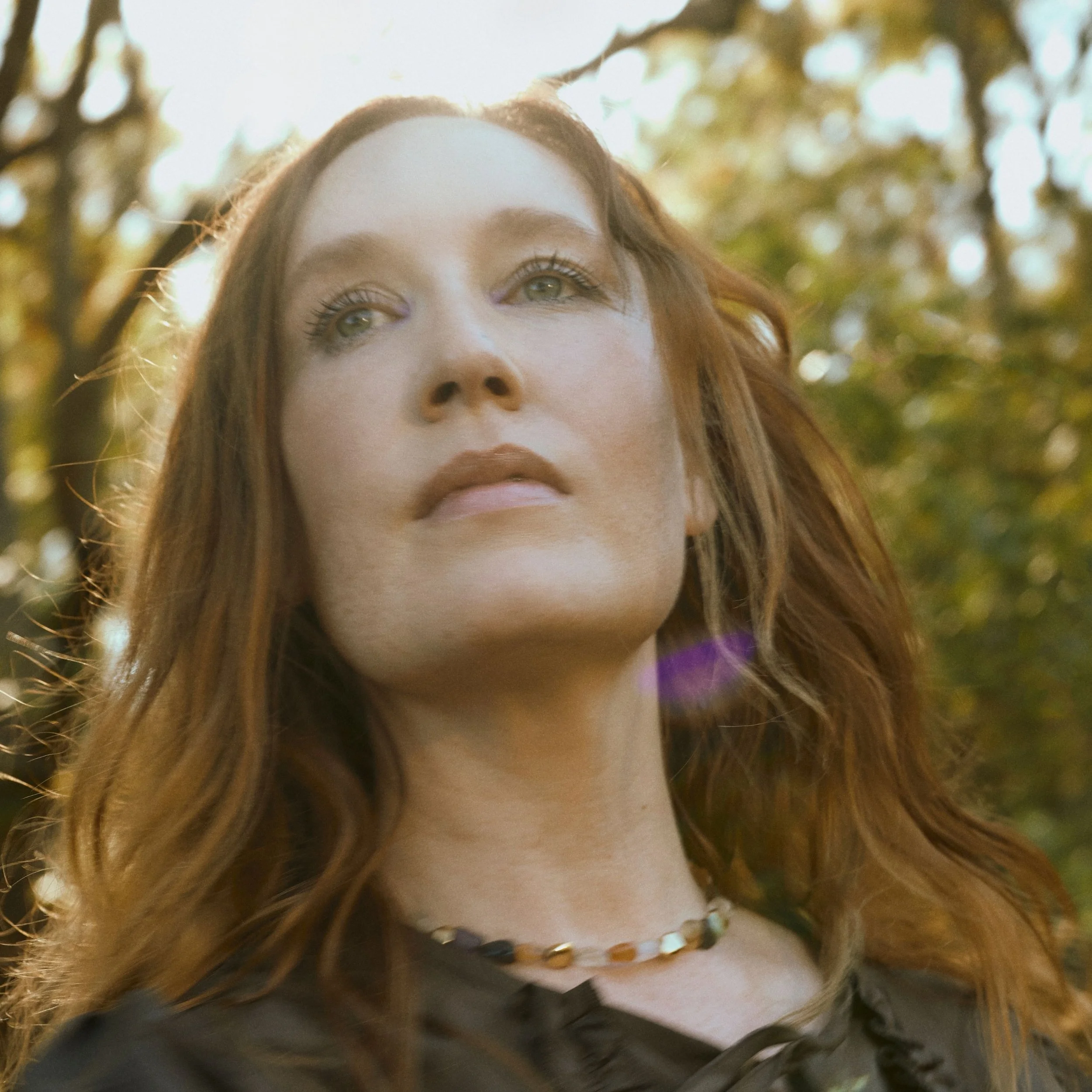A woman with long, wavy red hair standing outdoors with sunlight filtering through trees in the background.