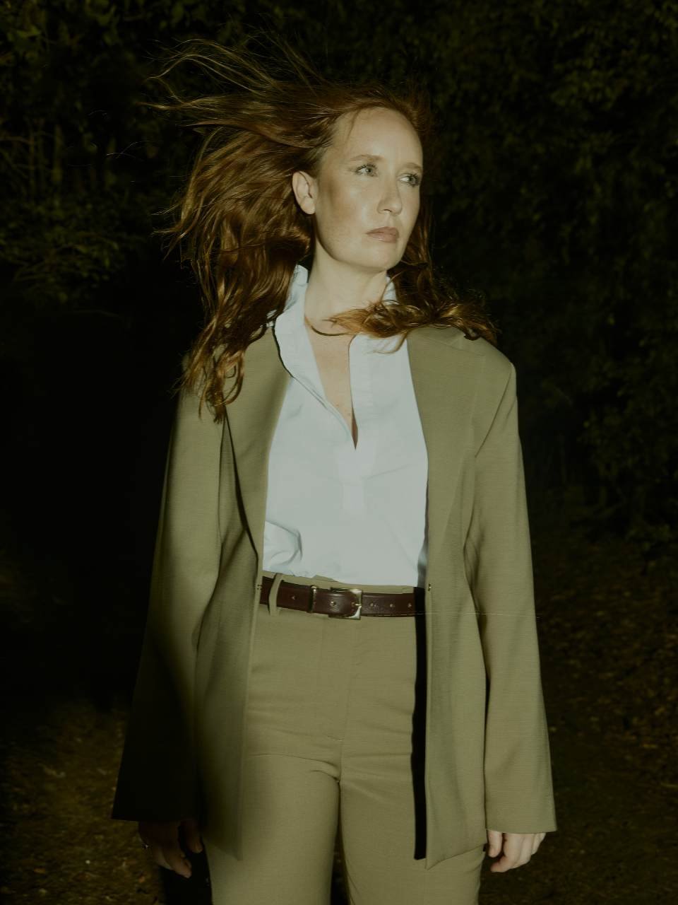 A woman with long red hair standing outdoors at night, wearing a tan suit and white shirt, with wind blowing her hair.