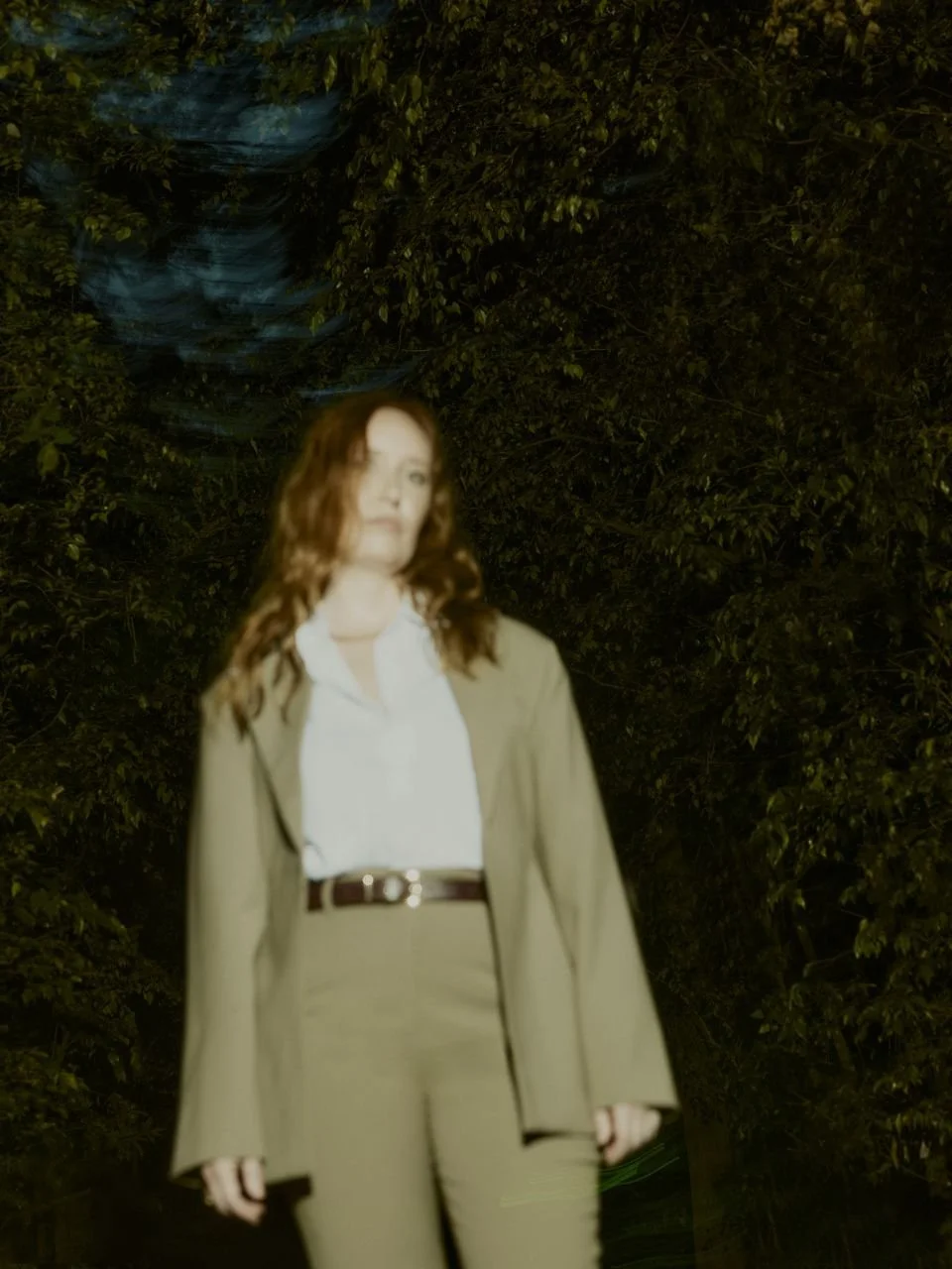 Woman with wavy hair standing outdoors at night, surrounded by trees, wearing a light-colored suit and white shirt.