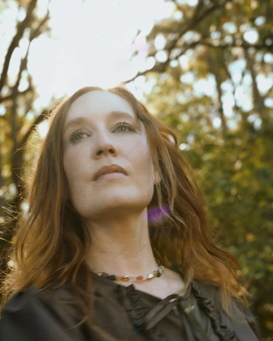 A woman with long, wavy red hair standing outdoors with sunlight filtering through trees in the background.